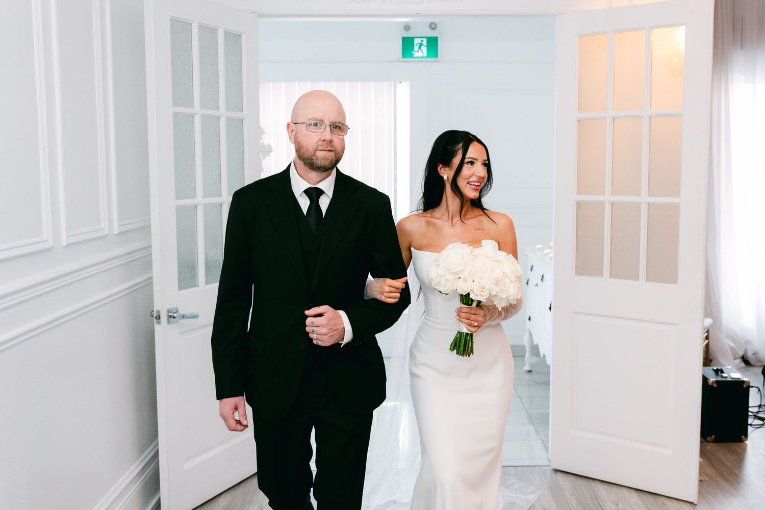 Wedding party members in formal black attire share emotional hugs and well-wishes in an elegant white venue.