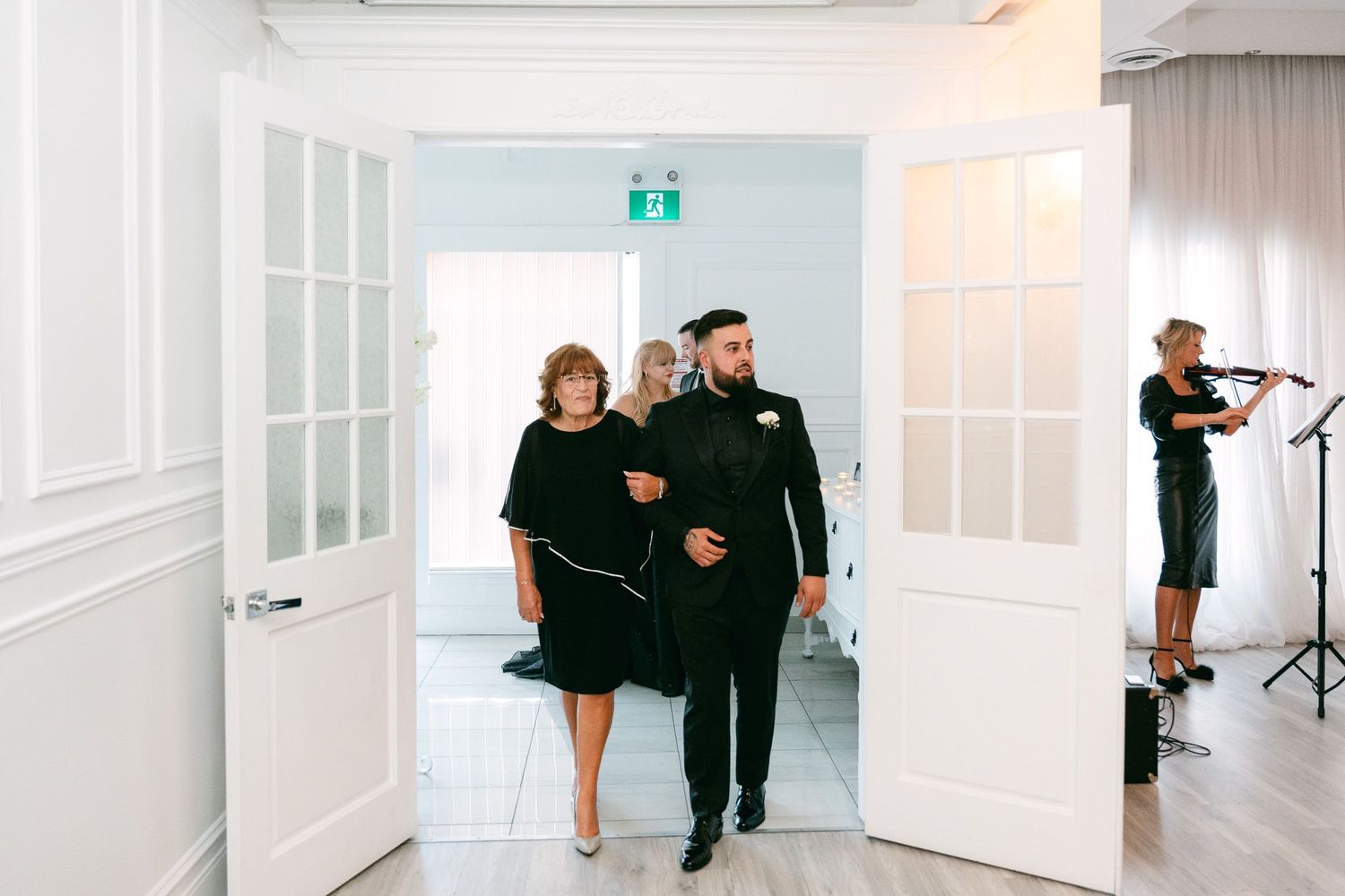 A couple walks through bright white elegant hallway doors while a violinist plays in the background.