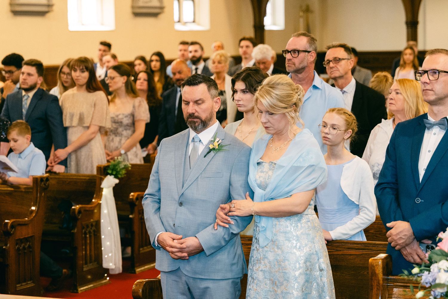 Groom in light blue suit walks down church aisle with elegant woman in lace dress during wedding ceremony.