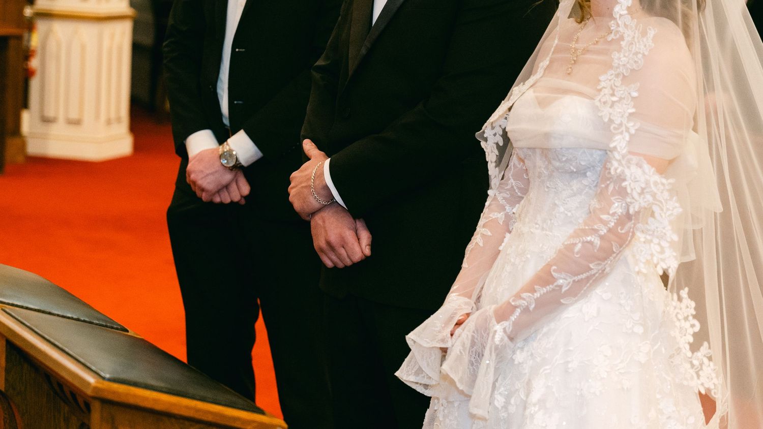 Bride in white lace wedding gown and veil stands at altar with groom in black tuxedo during church ceremony.