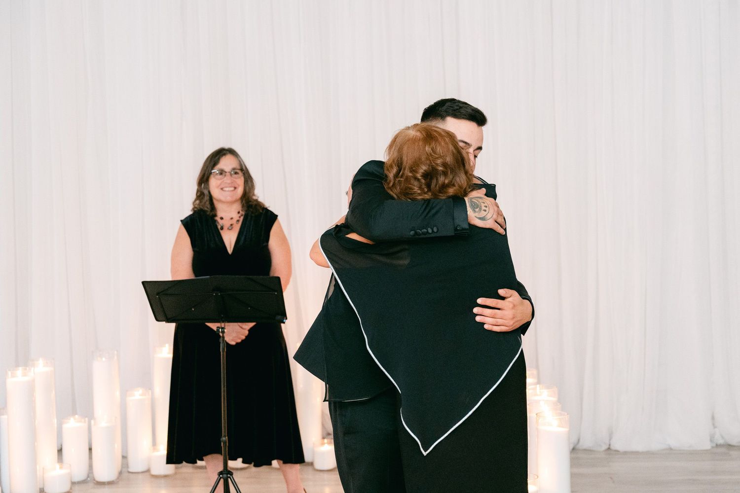 Two people embrace in a warm hug during a ceremony while an officiant stands at a podium in front of white drapes.