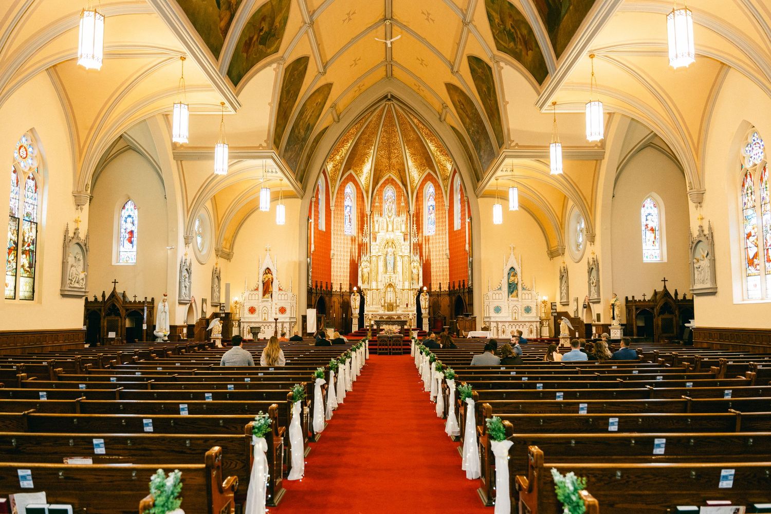 Elegant church interior with red carpet aisle, wooden pews, green floral decor, stained glass windows, and ornate golden altar.
