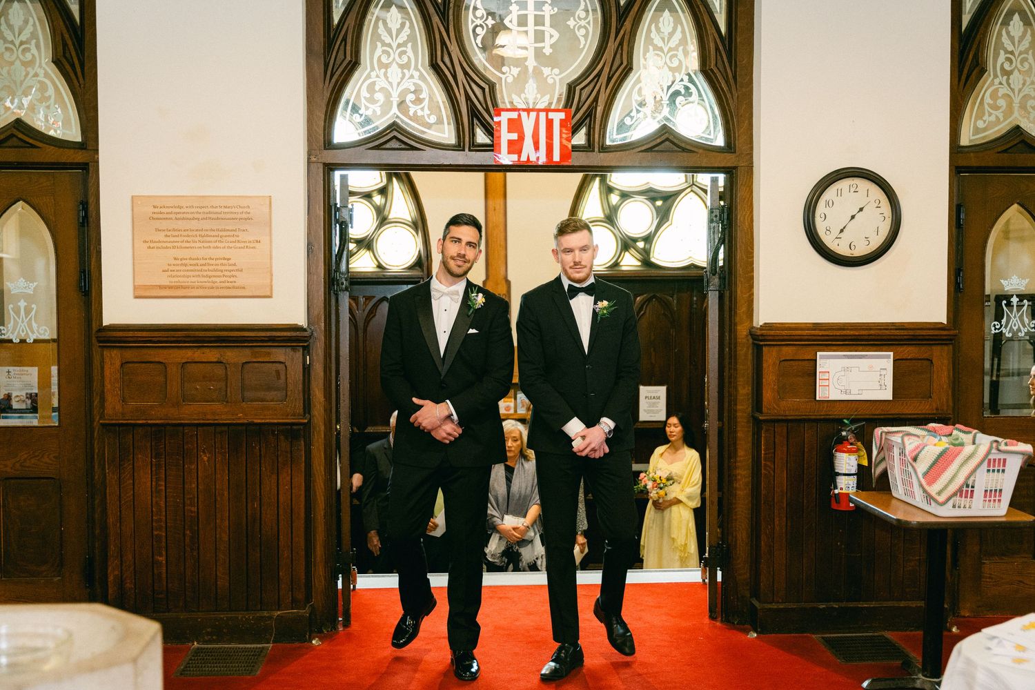 Two grooms in black tuxedos stand on a red carpet aisle inside an elegant church before their wedding ceremony.