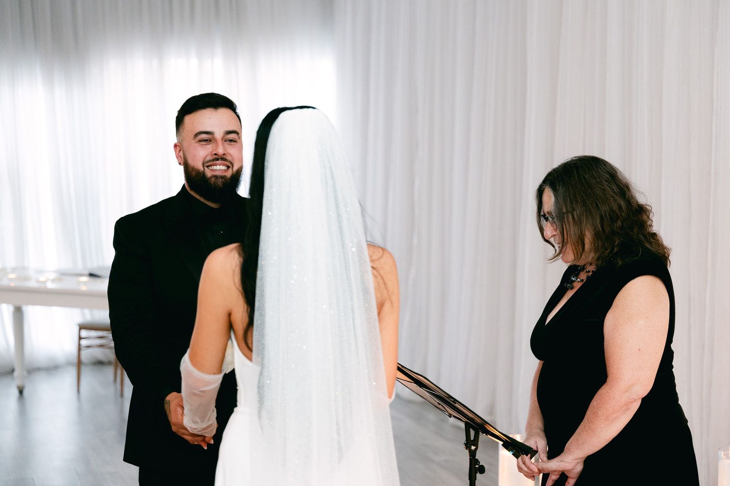 Wedding guests in formal attire share embraces in front of white curtained backdrop.