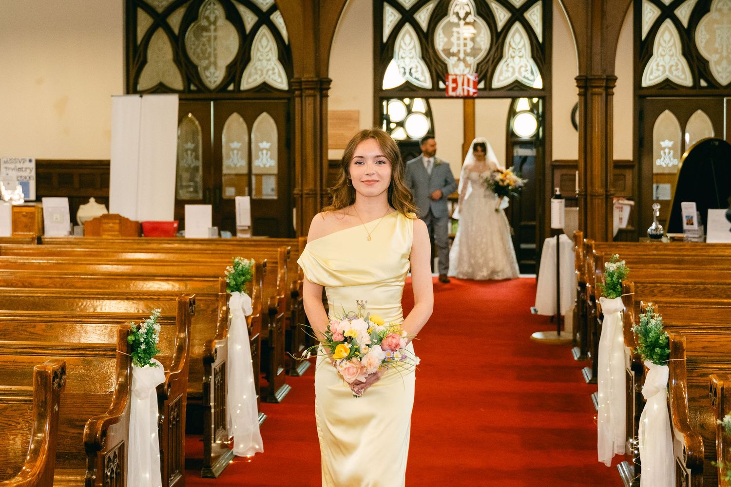Bridesmaid in gold dress holding bouquet stands in church aisle during wedding ceremony.