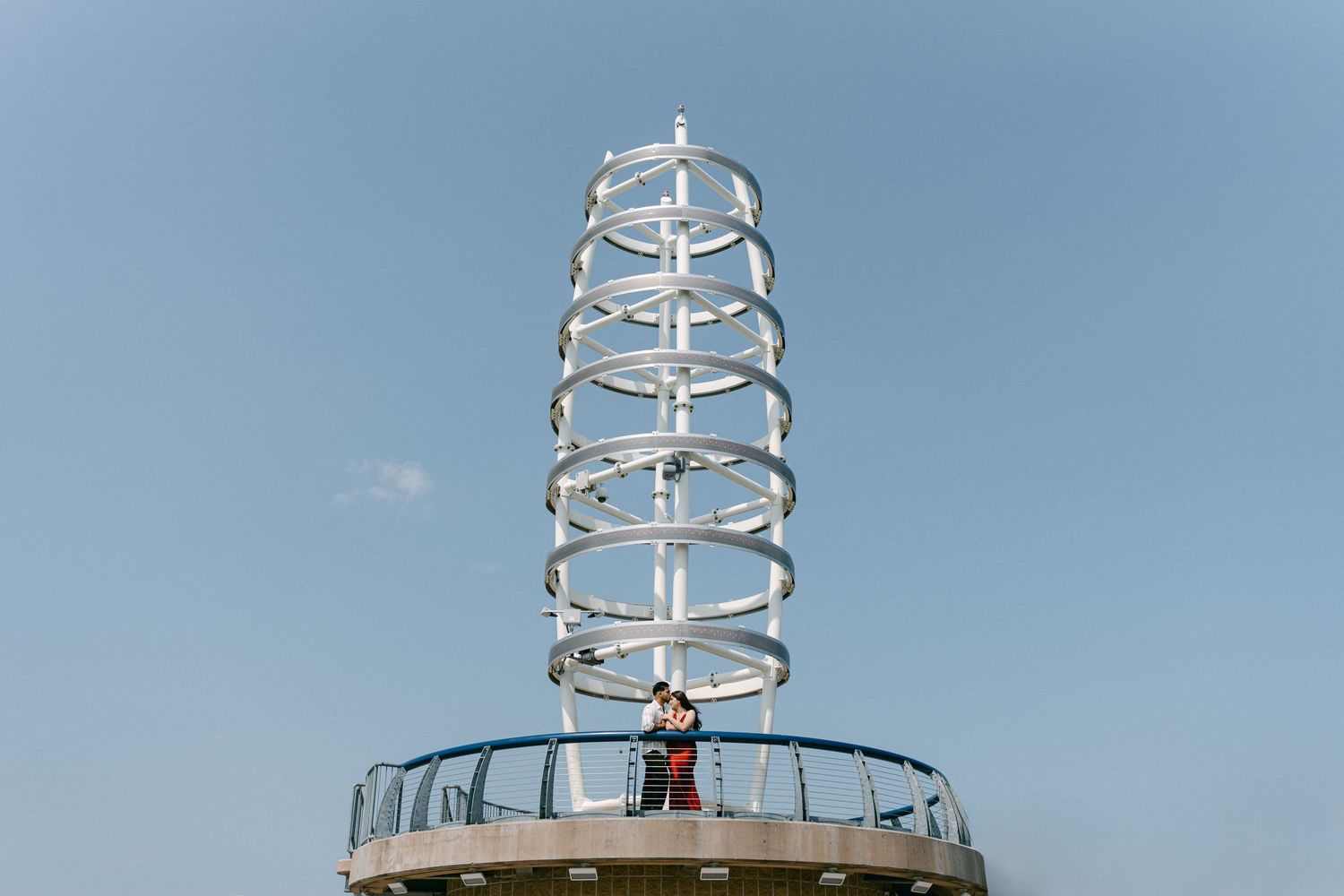 A couple stands atop a modern spiral tower structure against a blue sky.