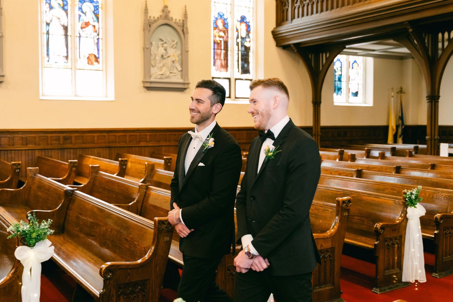 Two grooms in tuxedos smile at the altar inside a traditional wooden church during their wedding ceremony.