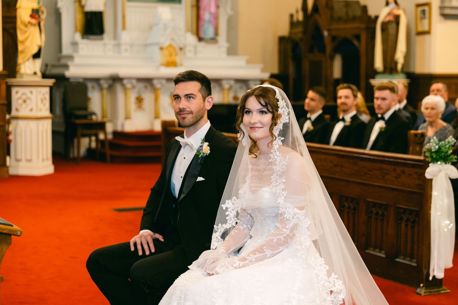 Bride and groom seated at church altar during wedding ceremony, bride wearing lace gown and veil, groom in black tuxedo.