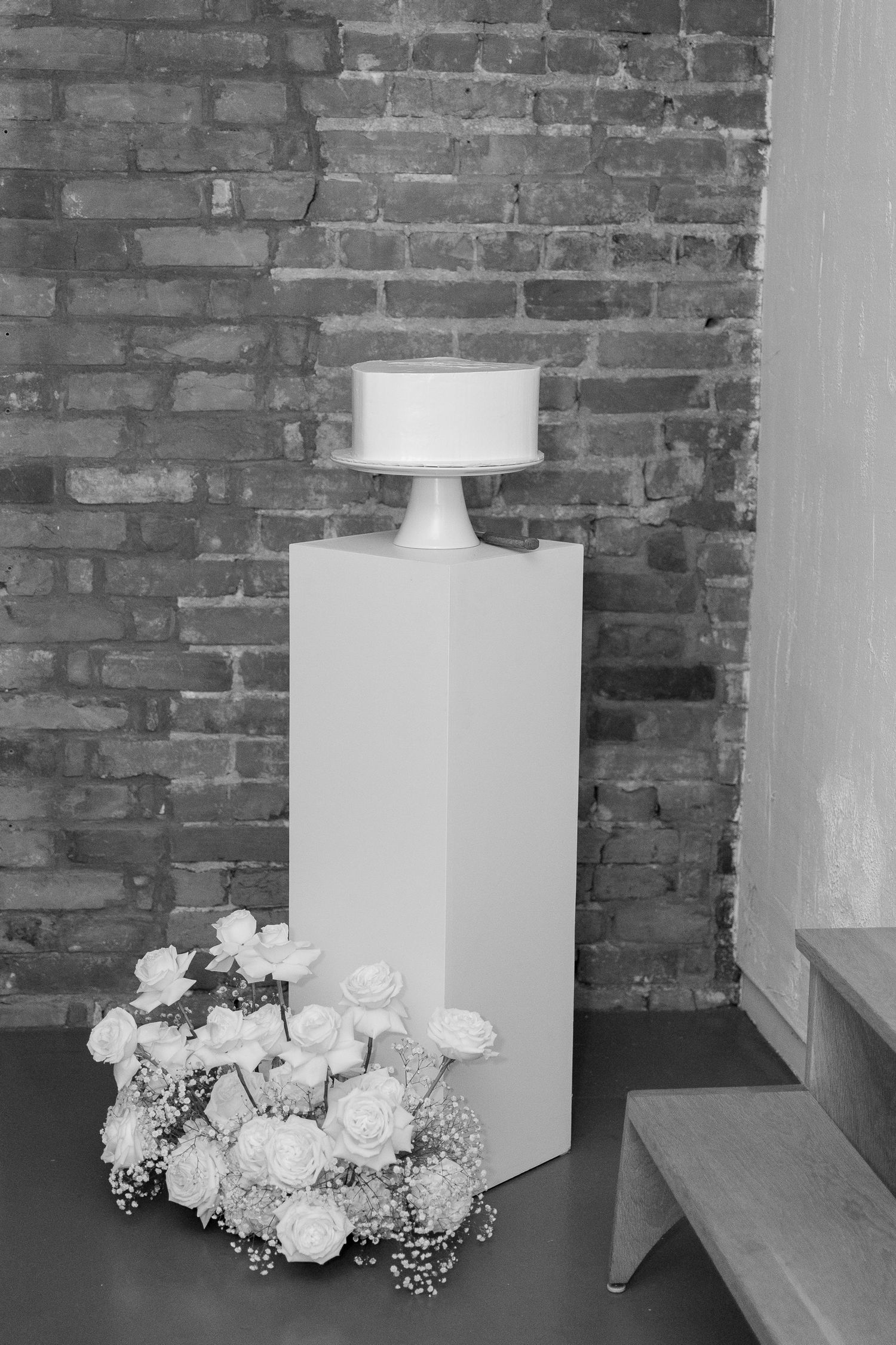 White wedding cake displayed on pedestal with fresh roses and baby's breath against rustic brick wall.