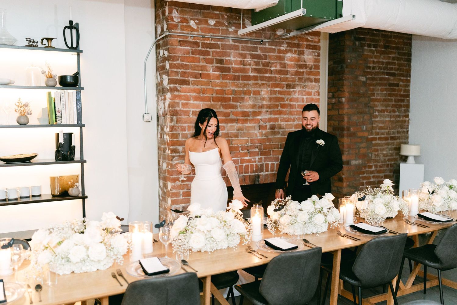 A formal celebration setup featuring elegant table settings and white floral arrangements against exposed brick walls.