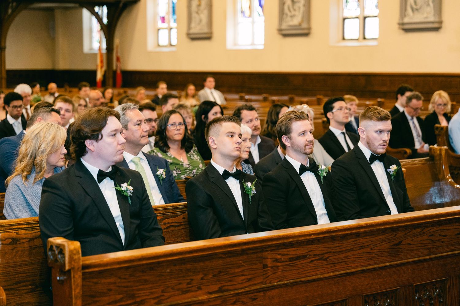 Groomsmen in black tuxedos with bow ties seated in church pews during a wedding ceremony.