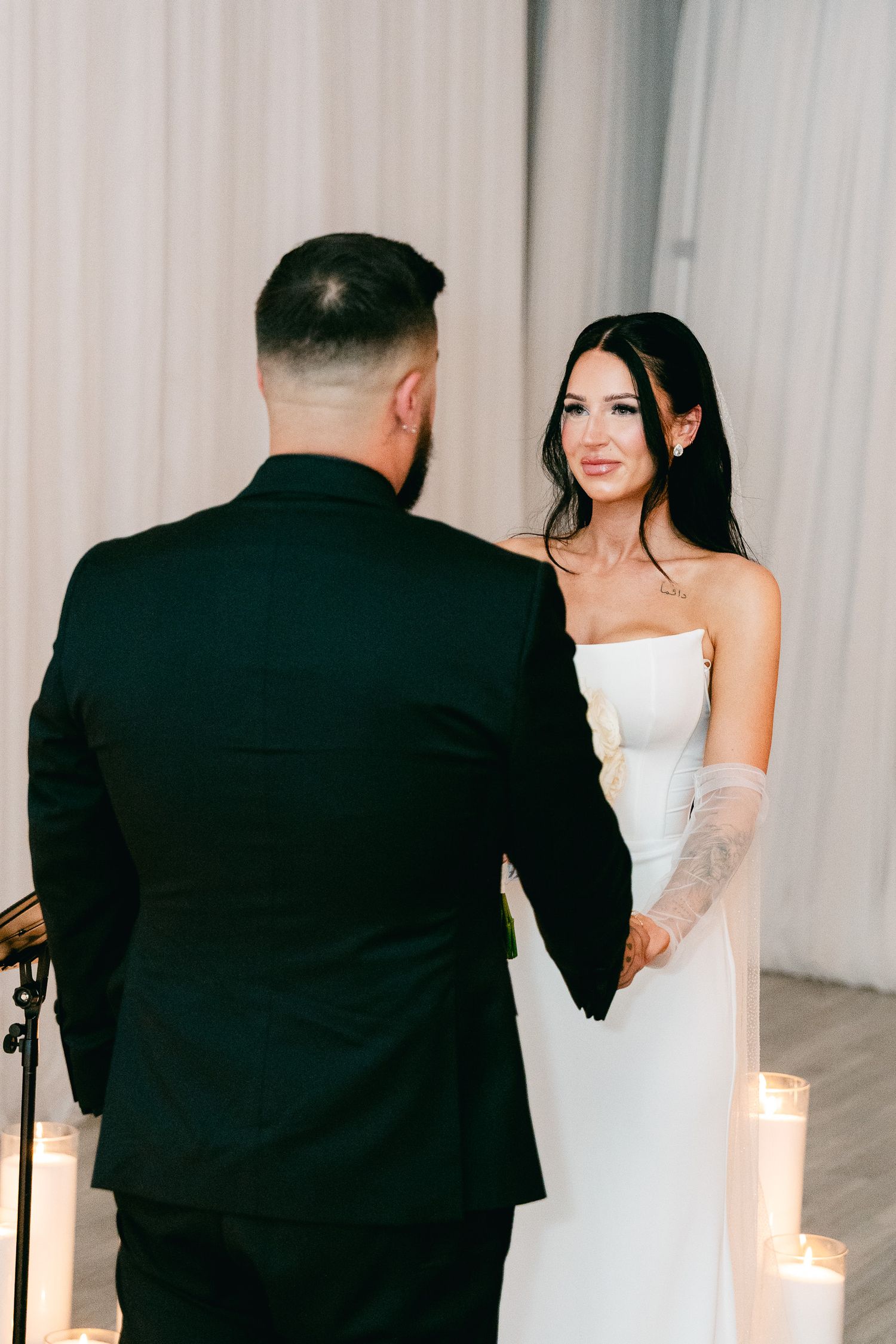 Bride in white strapless gown looks lovingly at groom during intimate wedding ceremony surrounded by candles.