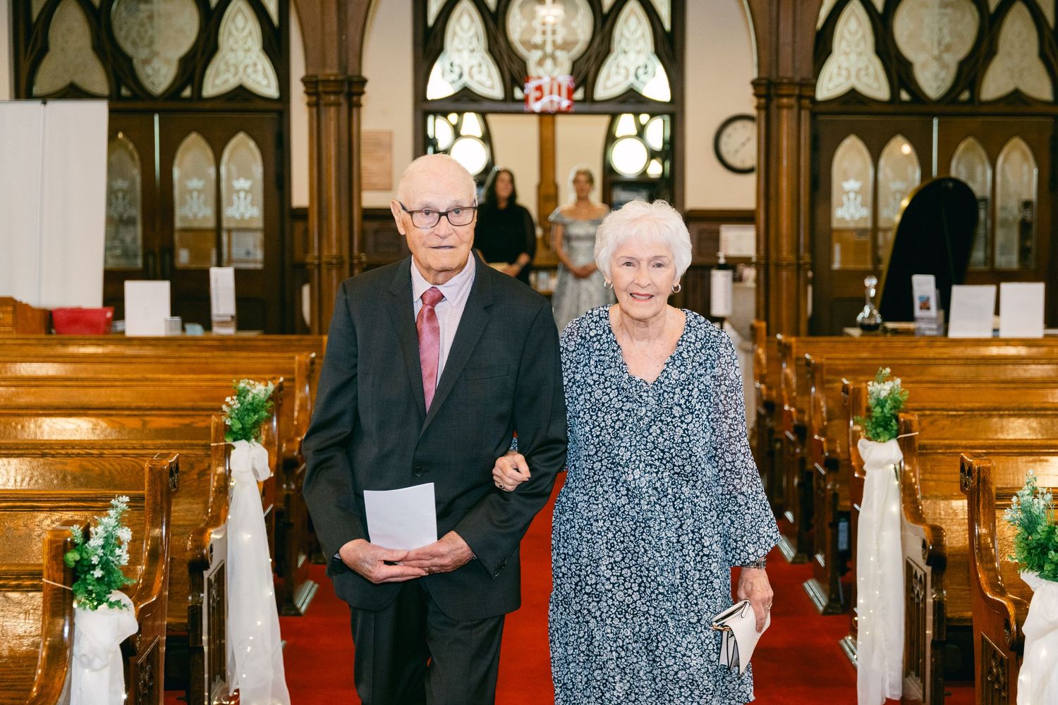 Elderly couple walking down church aisle during wedding ceremony, dressed in formal attire, surrounded by wooden pews.