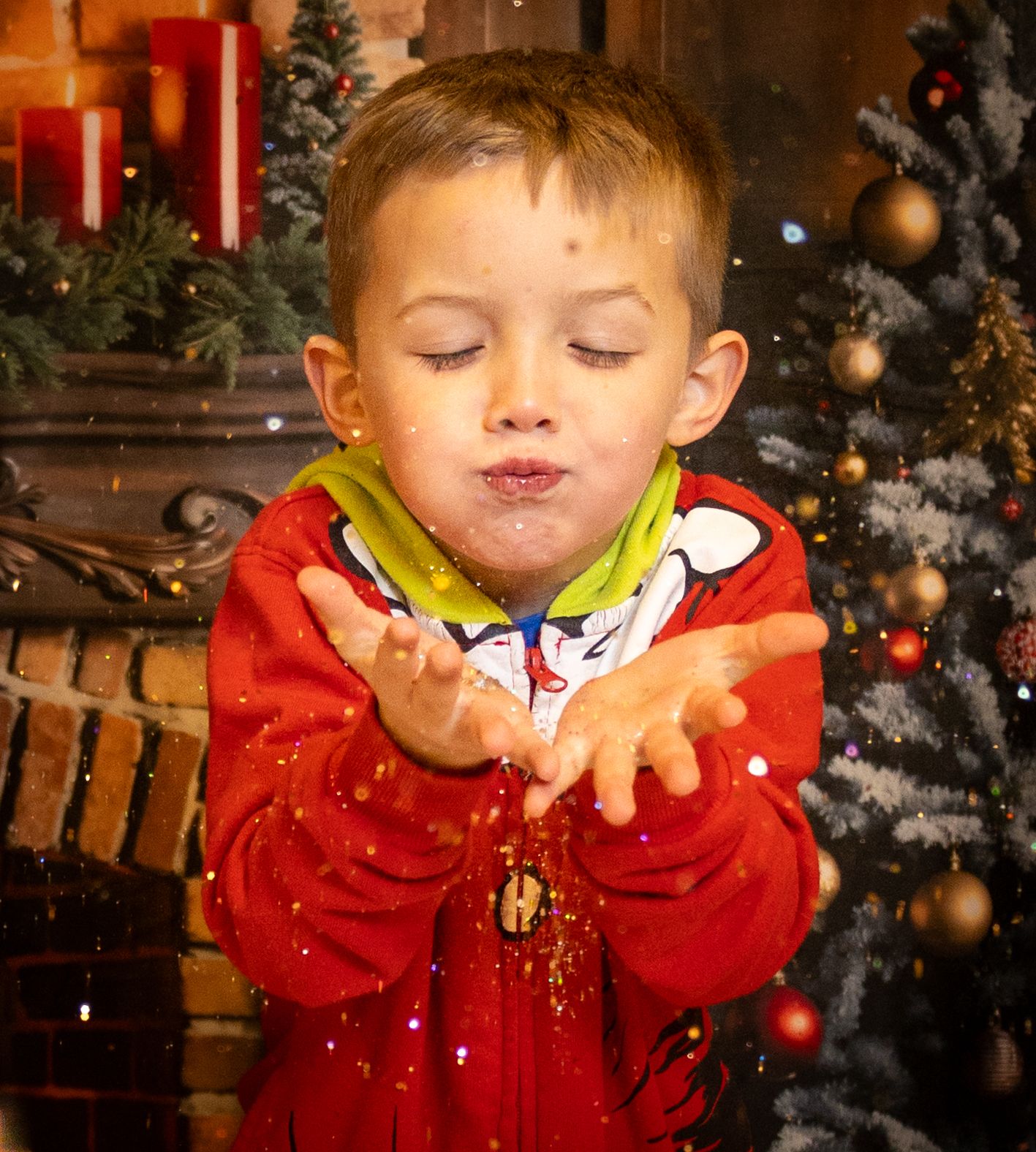 Young child in red jacket blows magical sparkly snow in front of decorated Christmas tree during holiday season.
