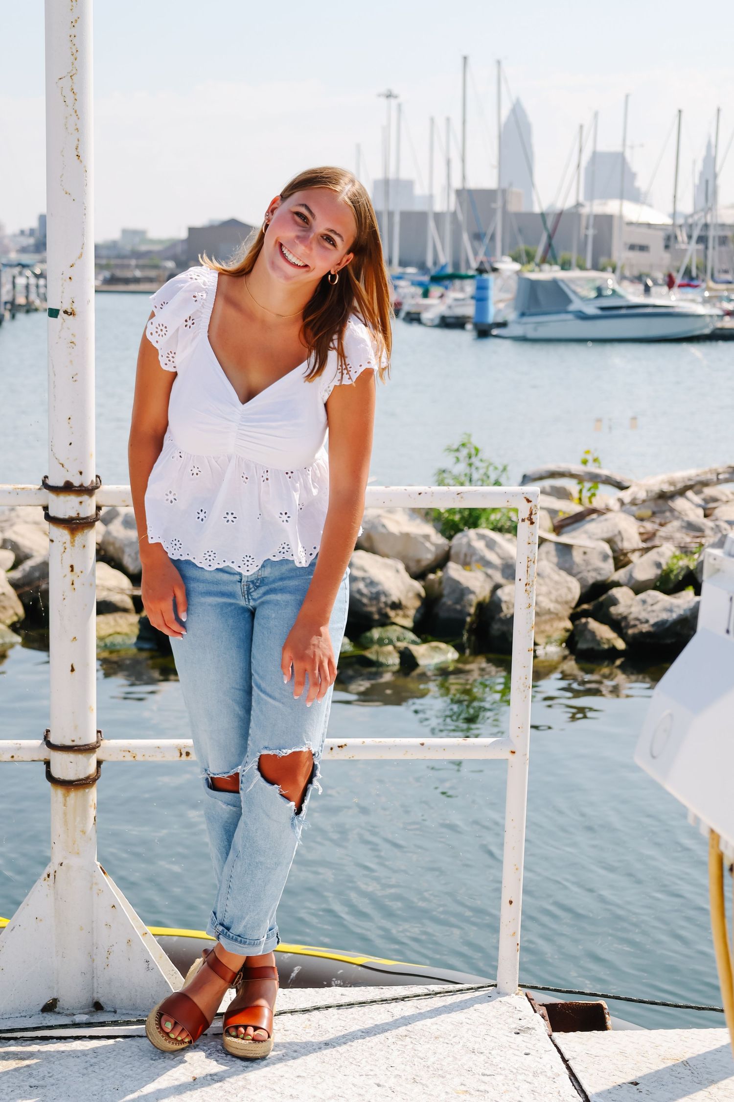 Woman in white eyelet top and ripped jeans poses by marina with boats and city skyline.