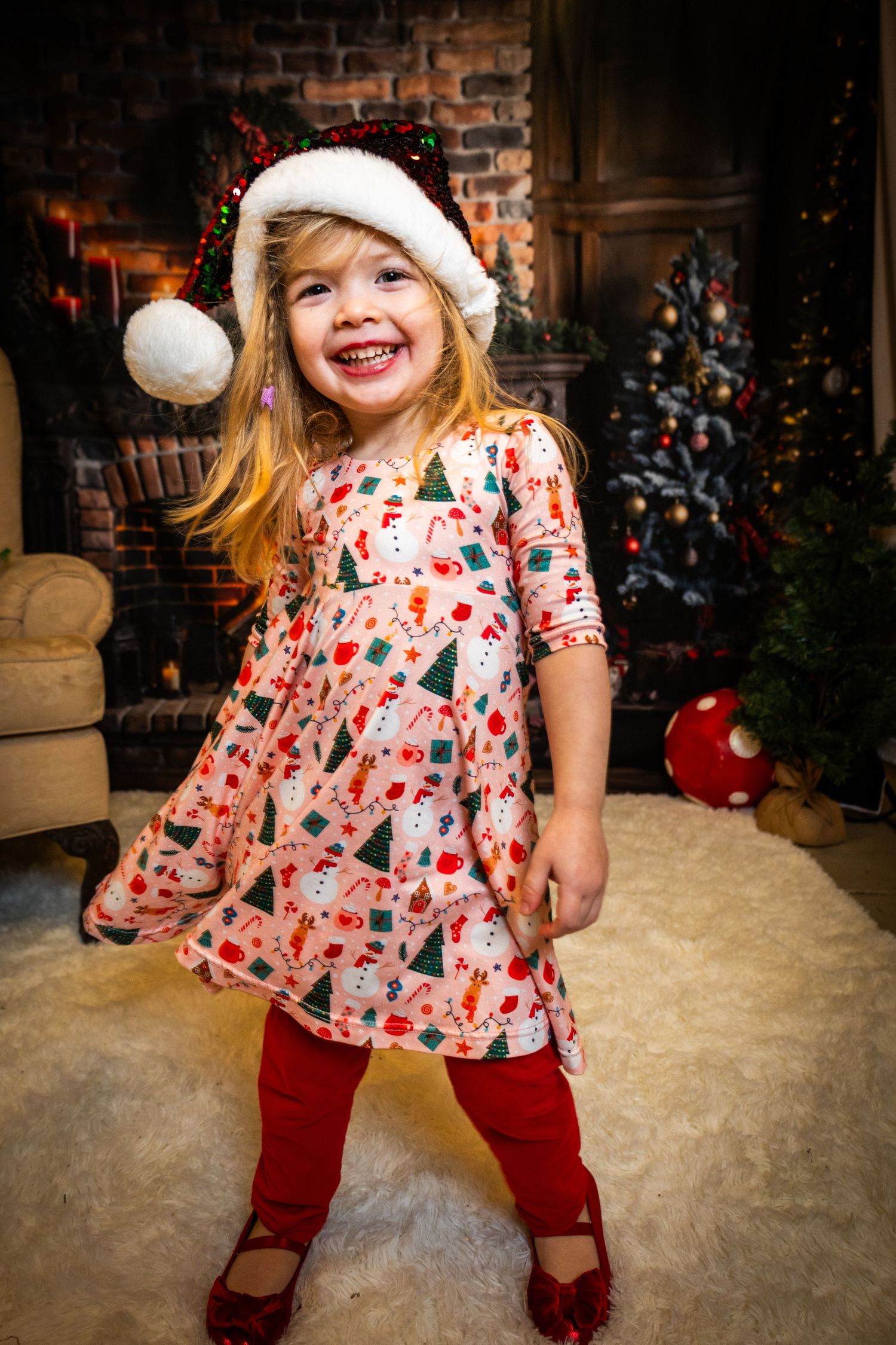 Young child in holiday attire with red pants and floral top dances playfully in front of brick fireplace during Christmas.