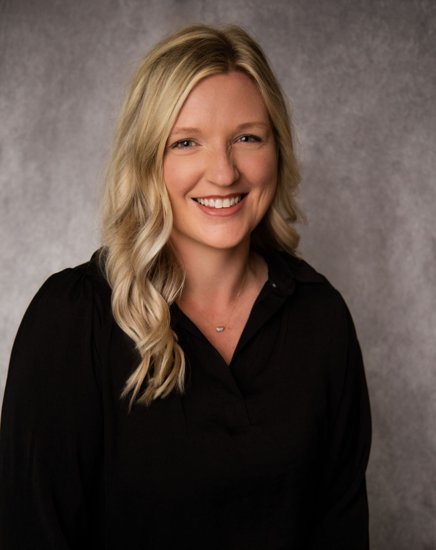 Professional headshot of a smiling blonde woman in a black blouse against a gray studio background.