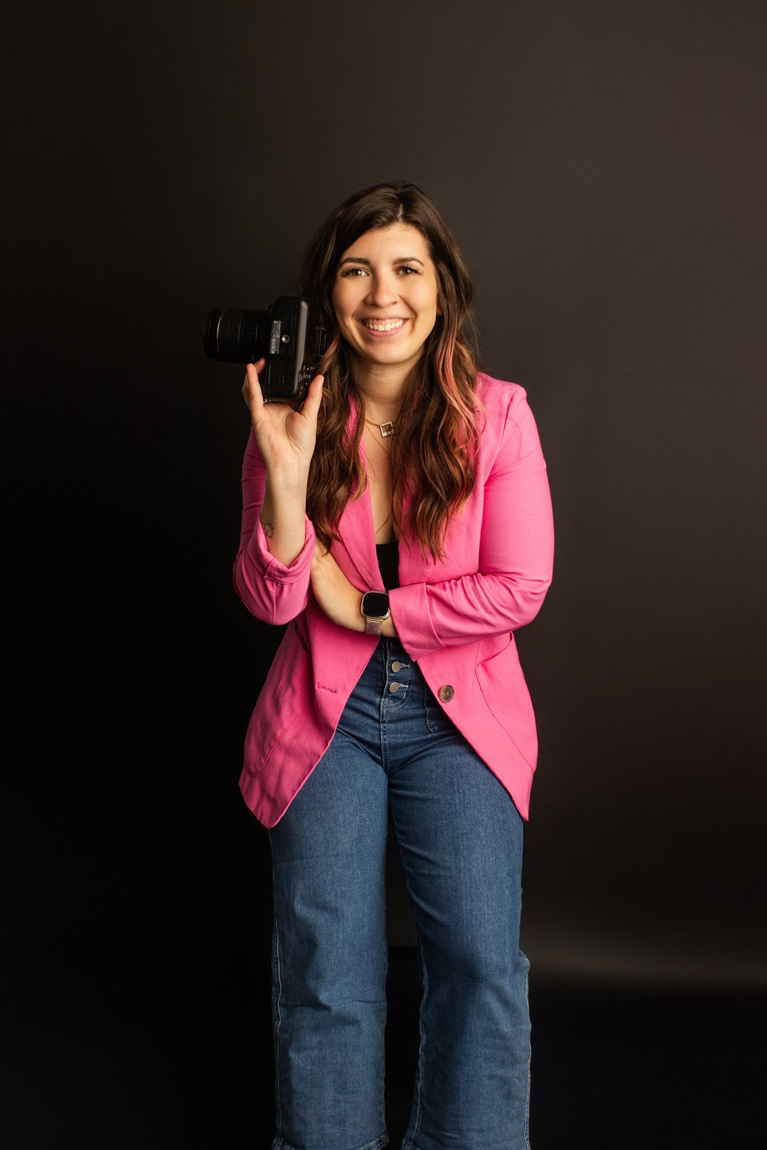 Woman in pink blazer and jeans smiling while holding a professional camera against a dark background.