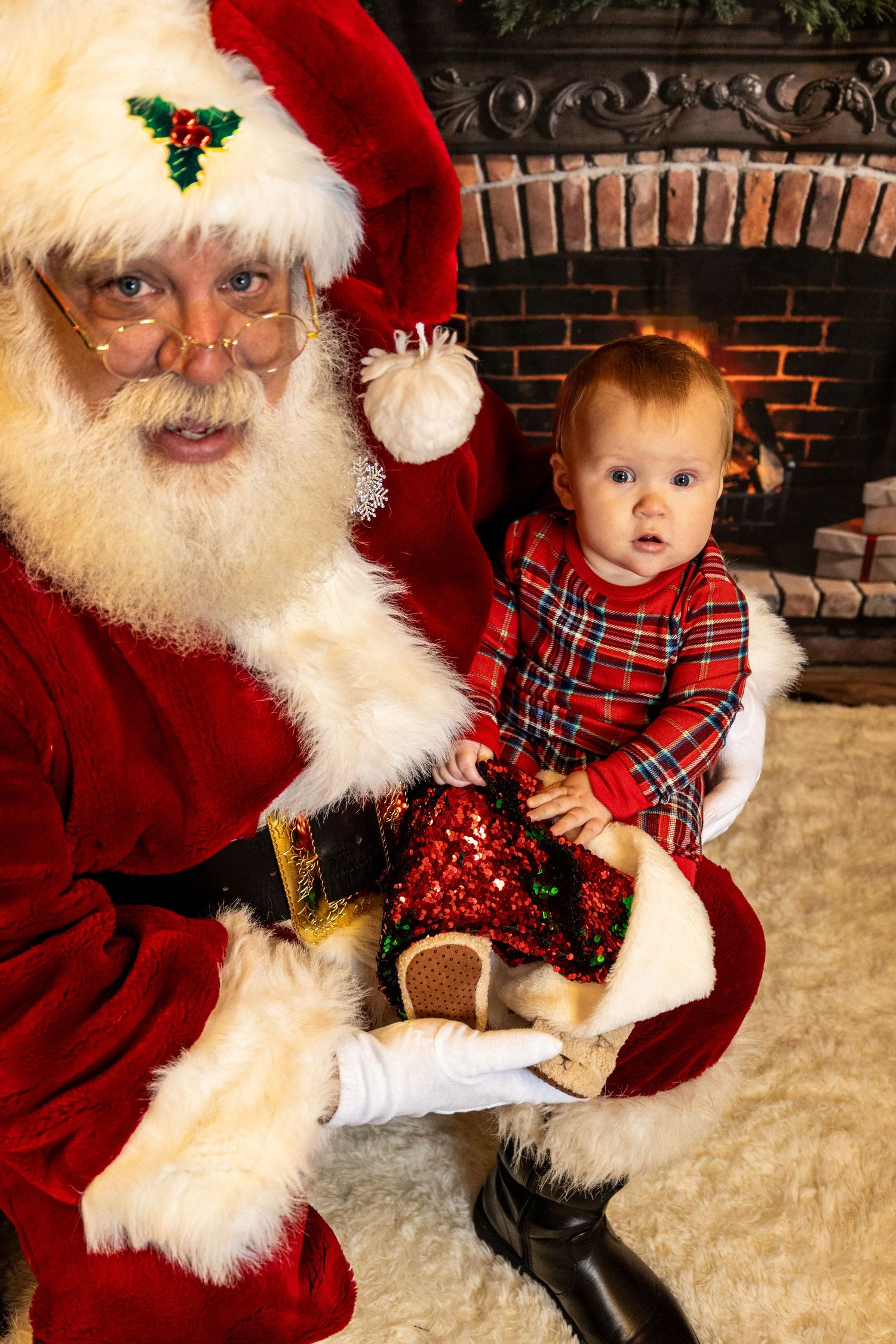 A small child in a red plaid dress sits on a traditional holiday figure's lap by a brick fireplace.