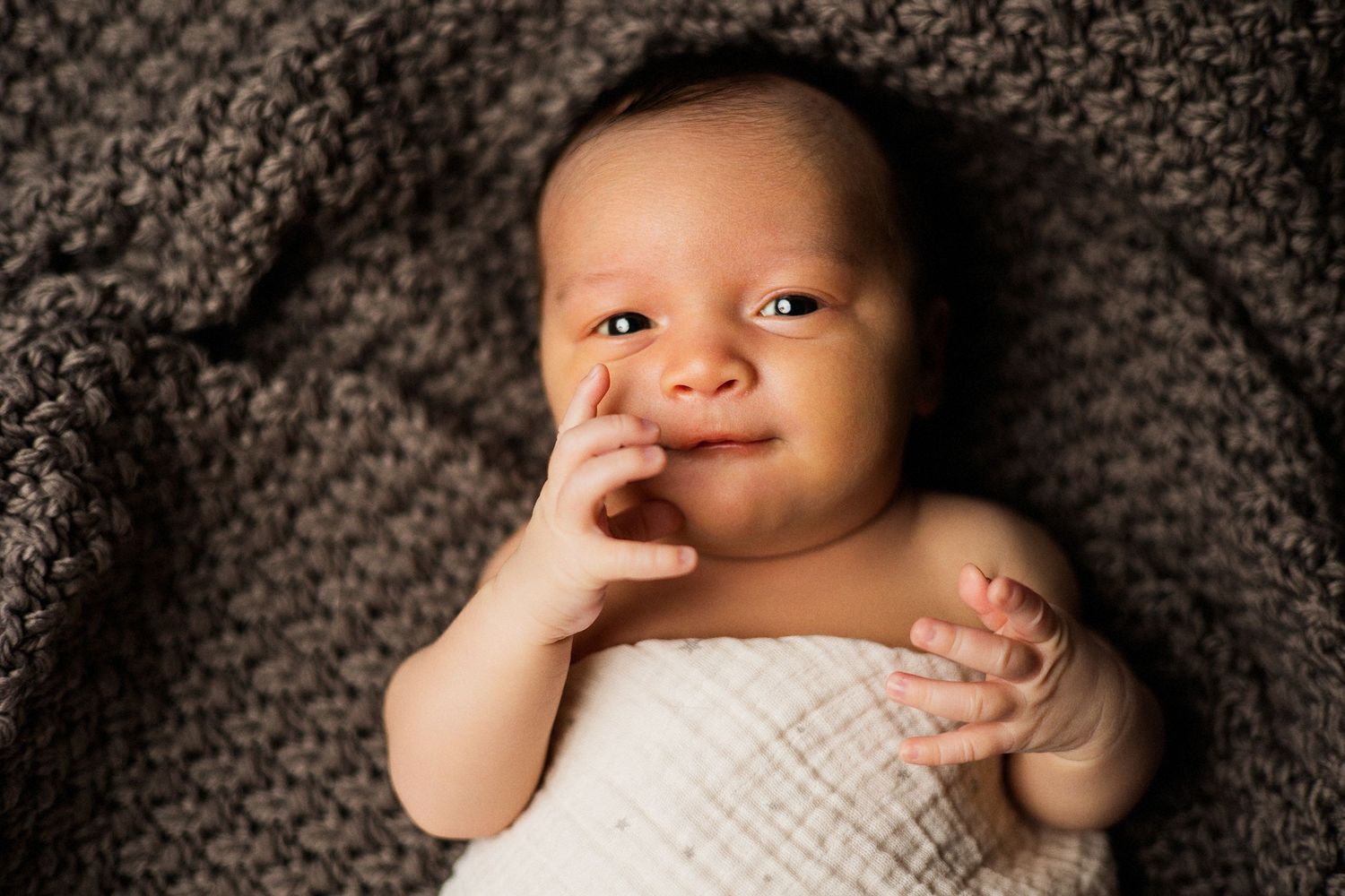 Adorable newborn baby smiling with finger near mouth, wrapped in white swaddle on a cozy brown knit blanket.