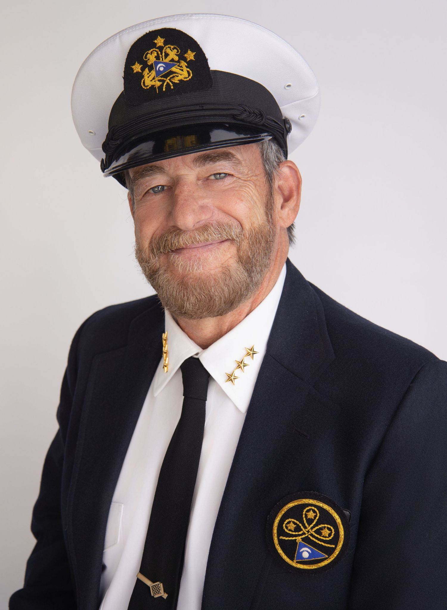 Man in naval captain uniform with white hat, dark blazer, tie, and gold insignia smiling for a portrait photo.