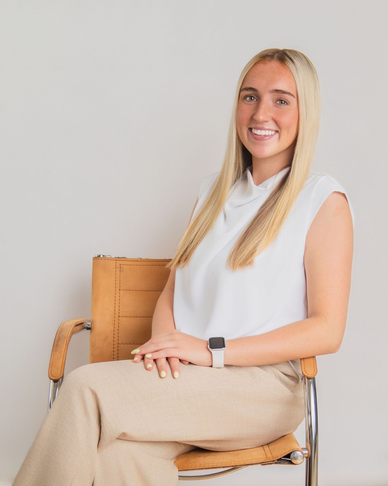 Smiling blonde woman in white sleeveless top and beige pants seated in a leather chair against a neutral background.