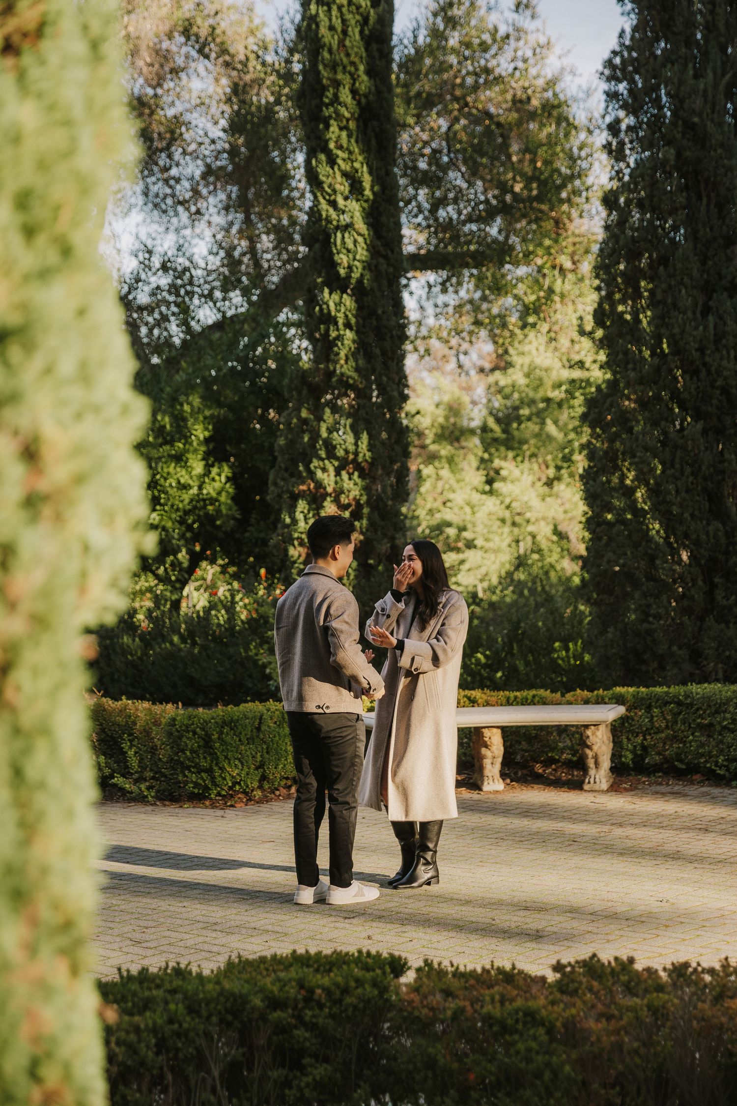Couple shares an intimate moment during outdoor engagement session in a lush garden setting with tall cypress trees.