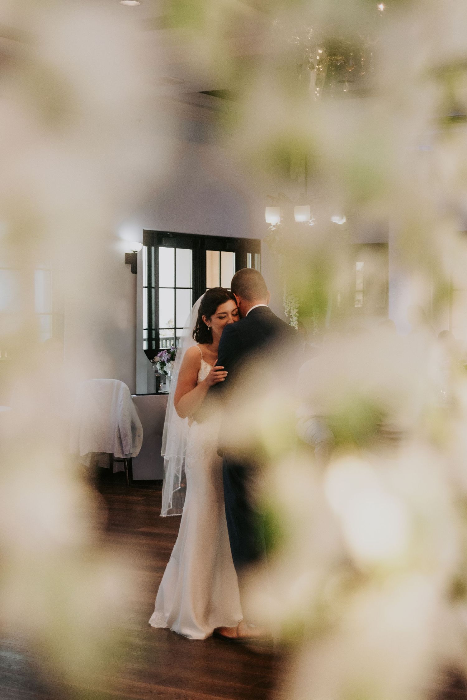 Romantic wedding dance captured through soft focus floral decorations in elegant indoor venue.