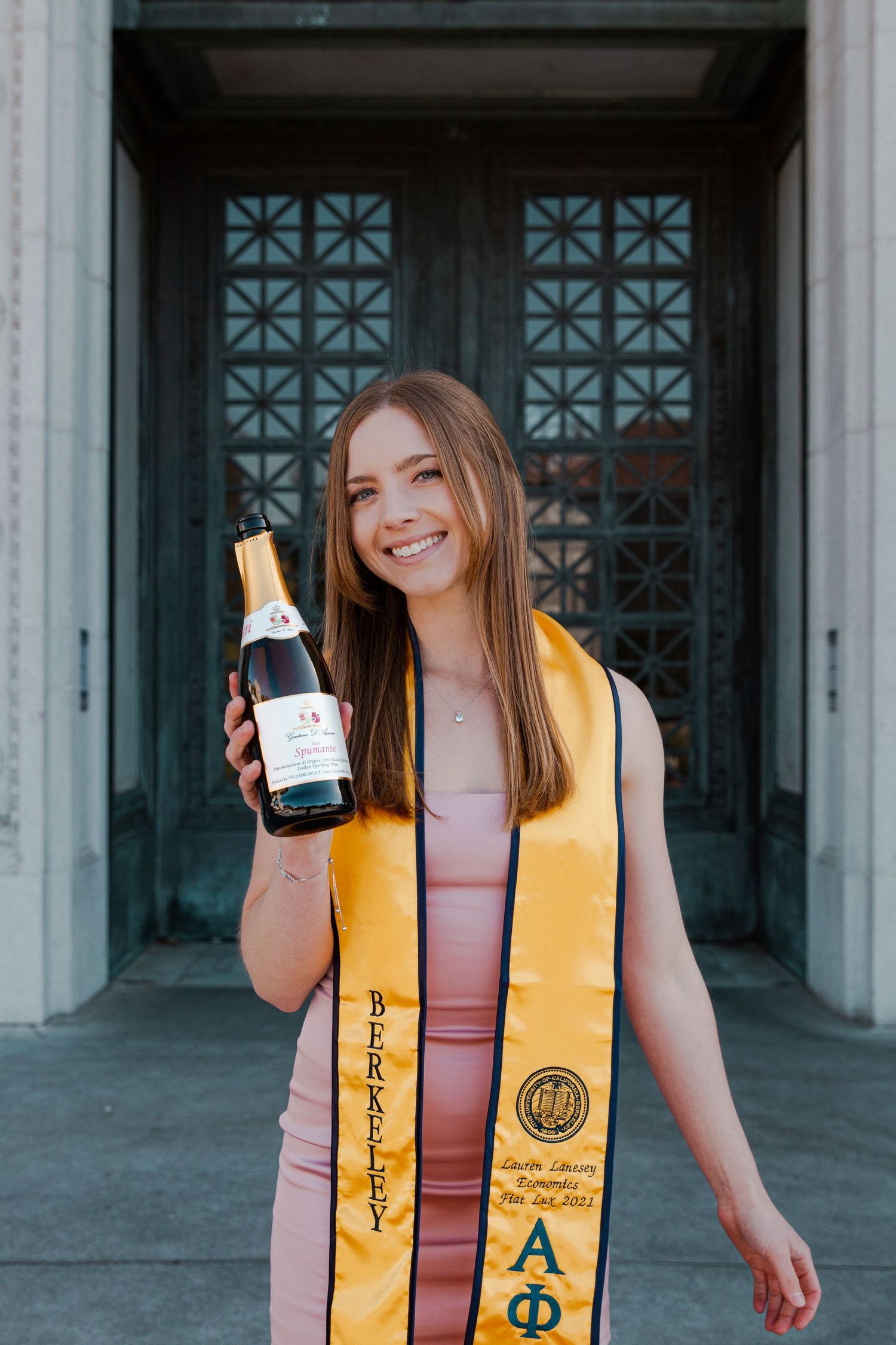 Graduate in yellow stole holds champagne bottle while posing in front of ornate doorway.