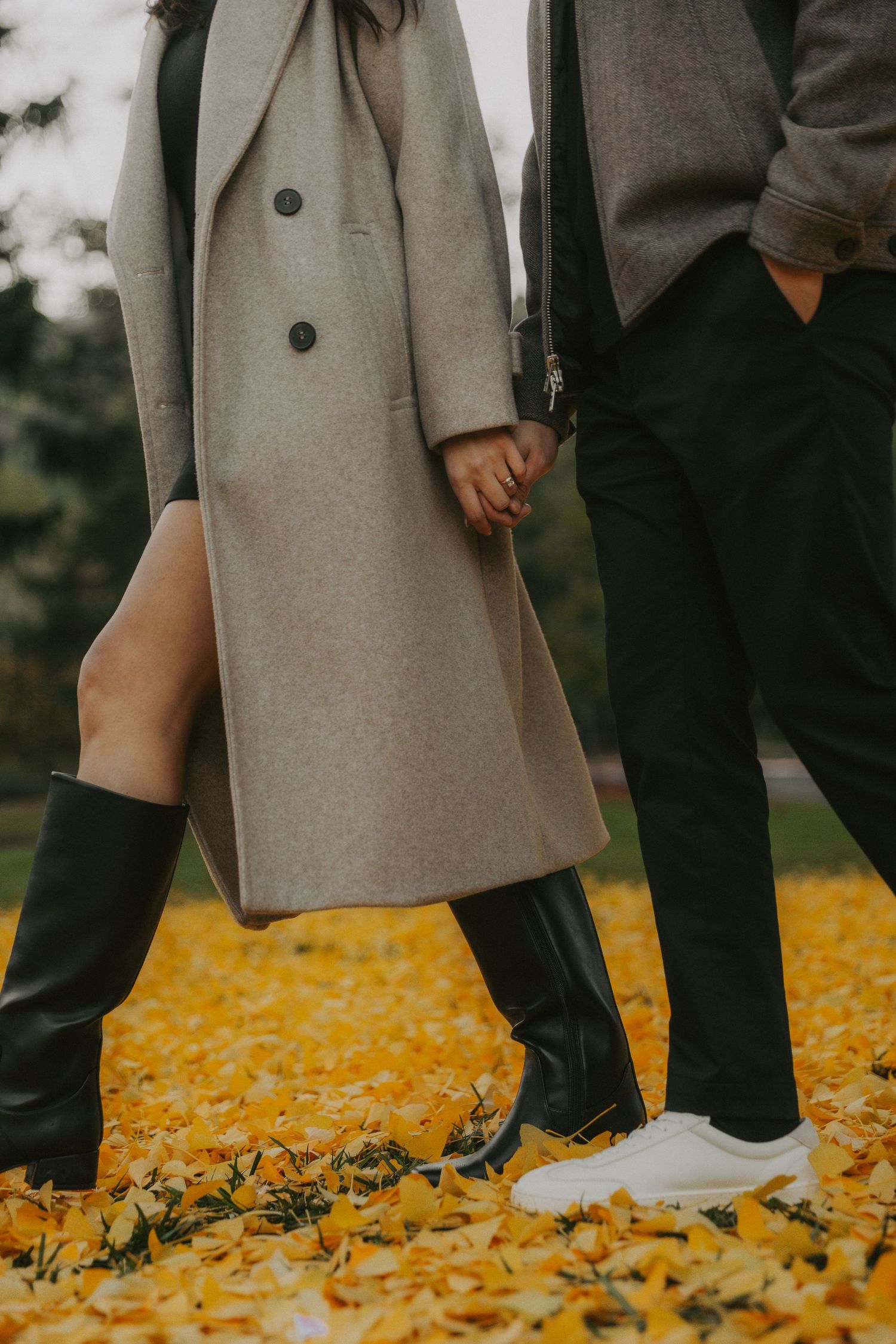 People wearing grey coats and black pants stand on vibrant yellow fallen autumn leaves in an outdoor setting.