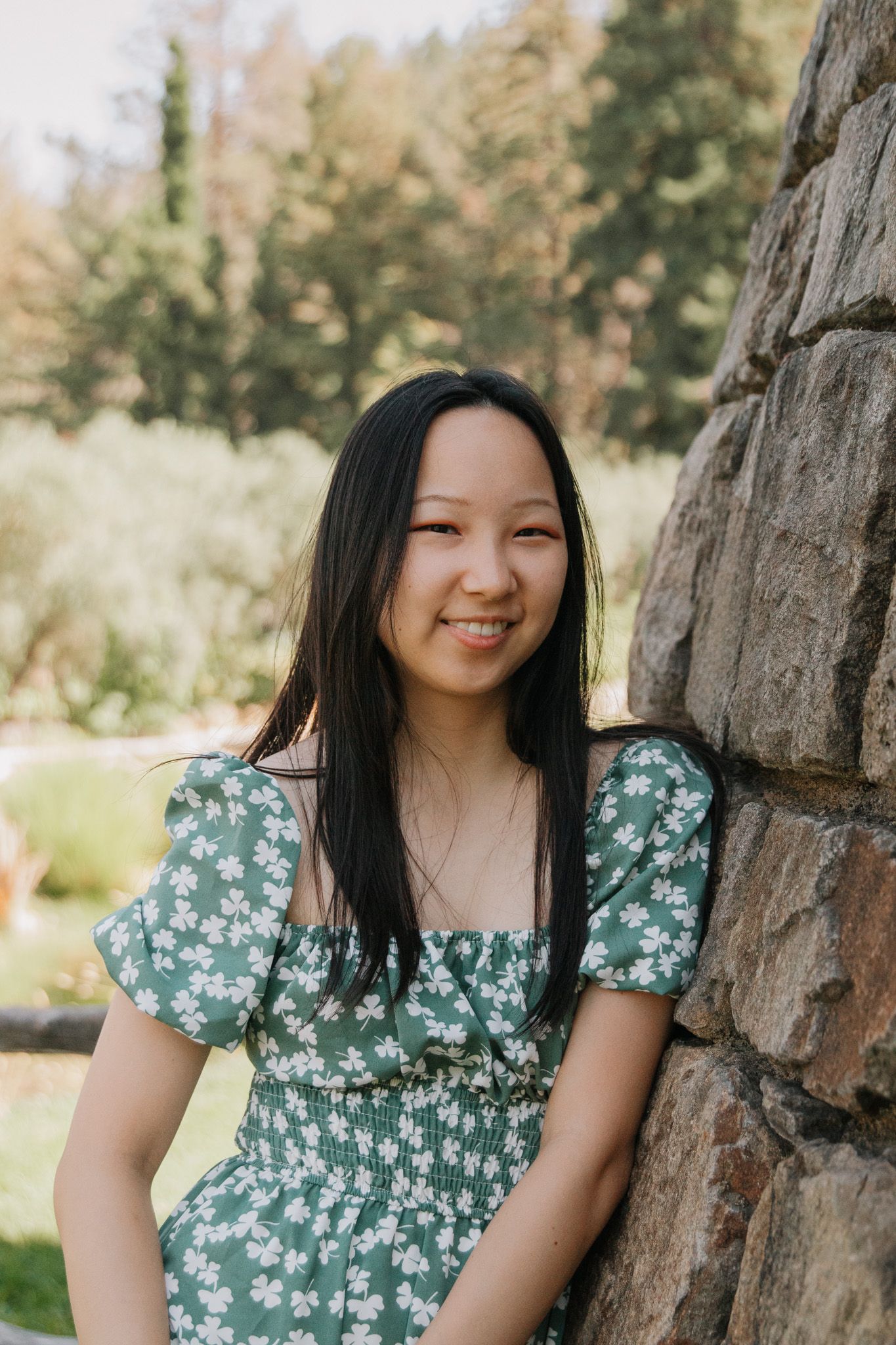 Person in a floral green dress smiling while leaning against a stone wall outdoors.