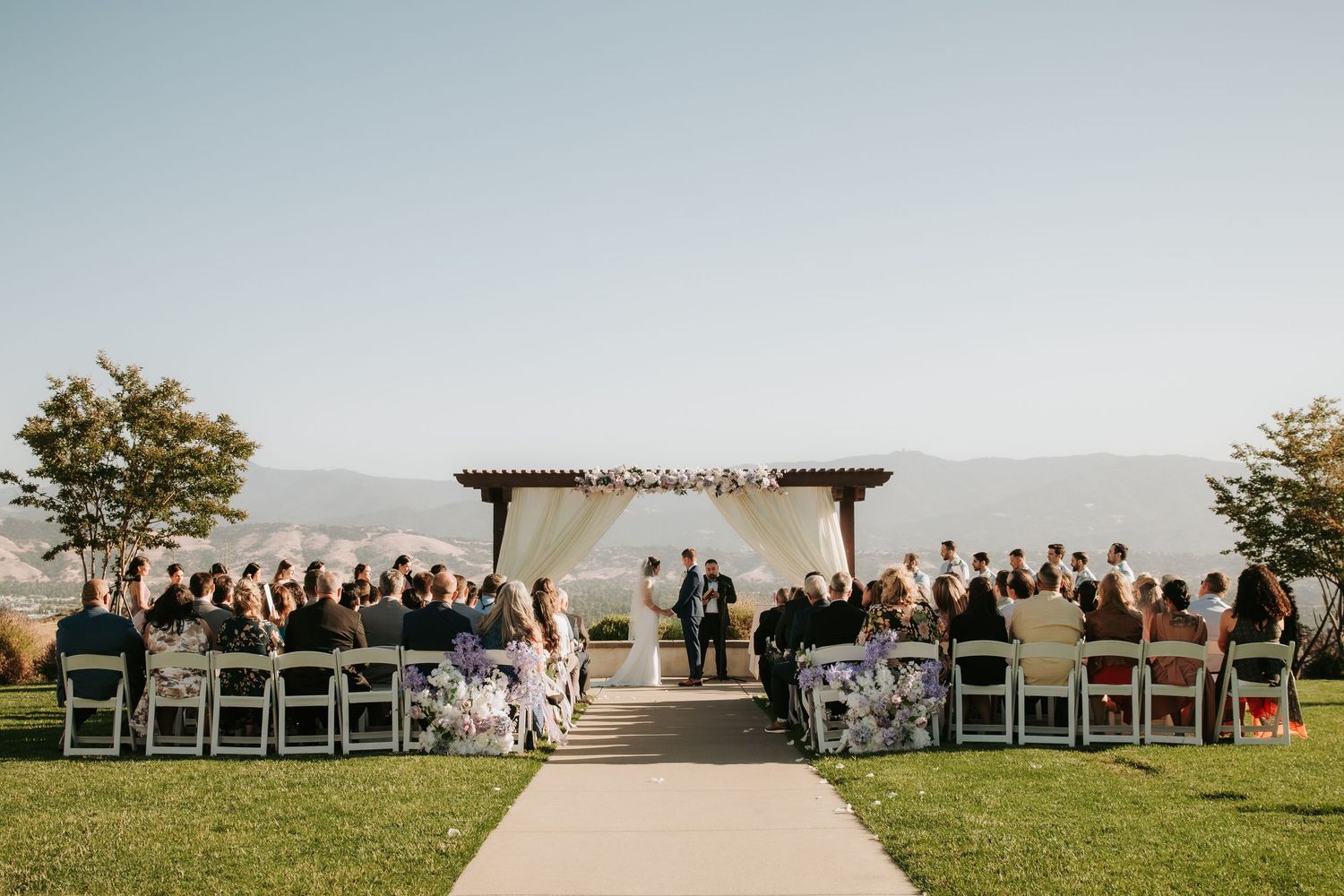 Outdoor wedding ceremony setup with white chairs and mountain backdrop at sunset.