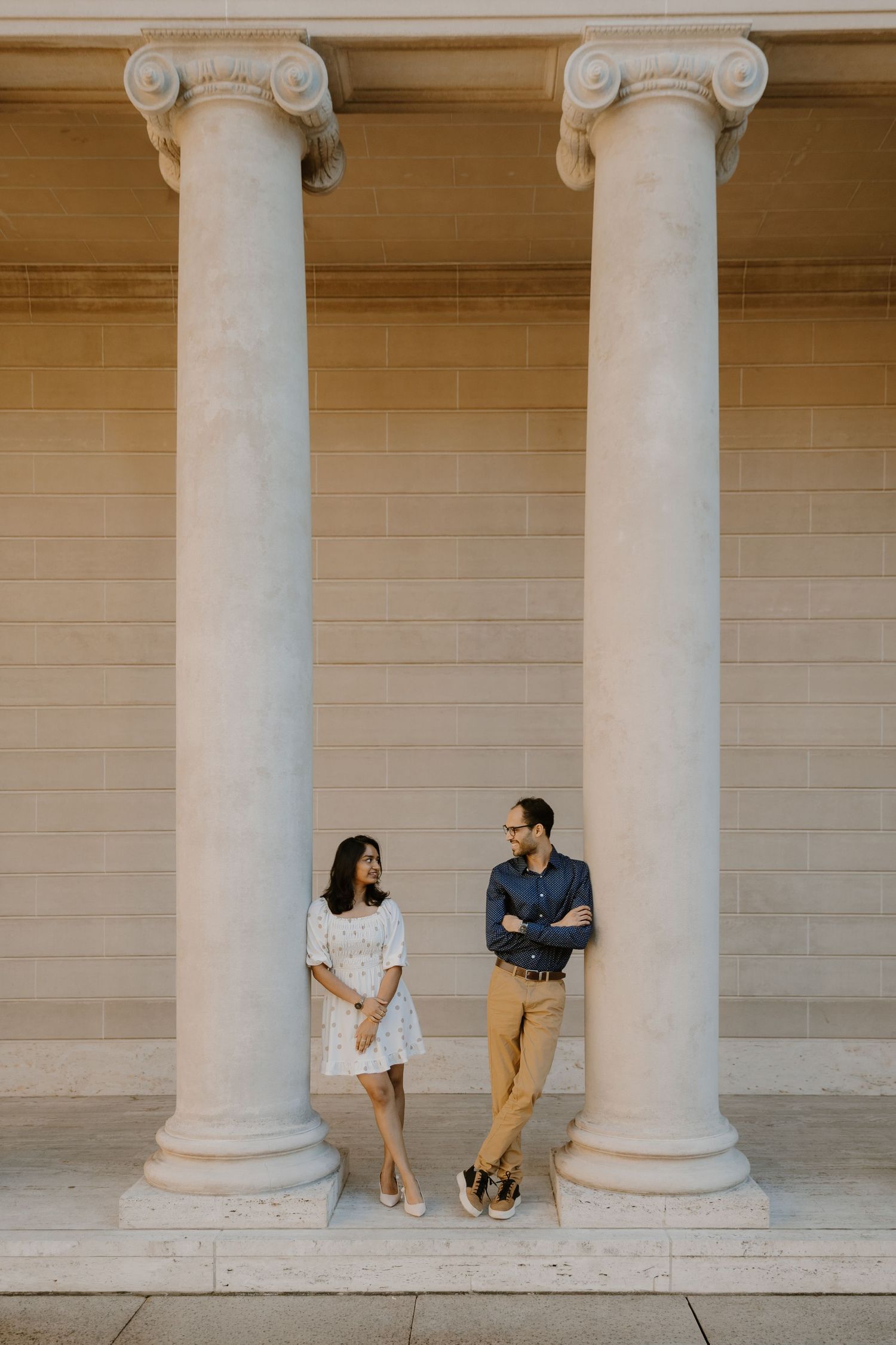 A couple sits together between elegant white columns in an architectural setting during a romantic photoshoot.