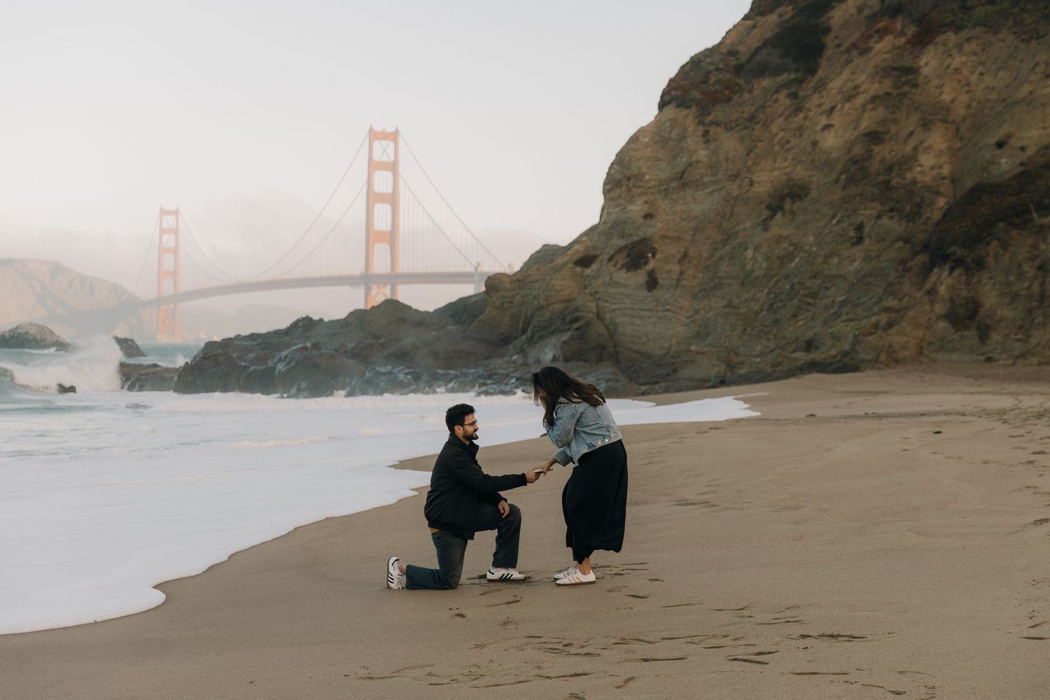 Two people sit on rocks at Baker Beach with views of the Golden Gate Bridge and San Francisco Bay at sunset.