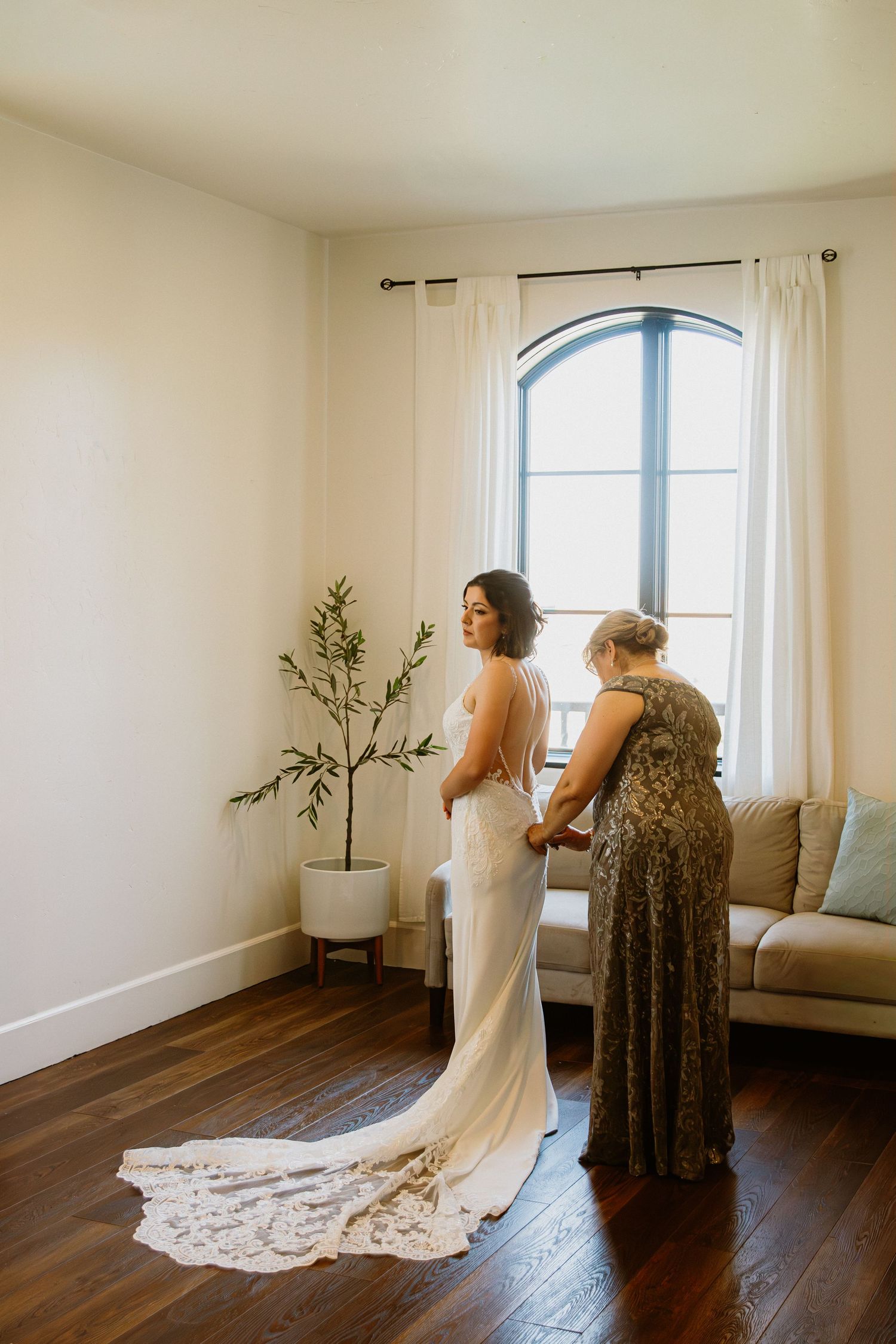 A wedding dress fitting scene in a bright room with hardwood floors, white walls and an arched window.