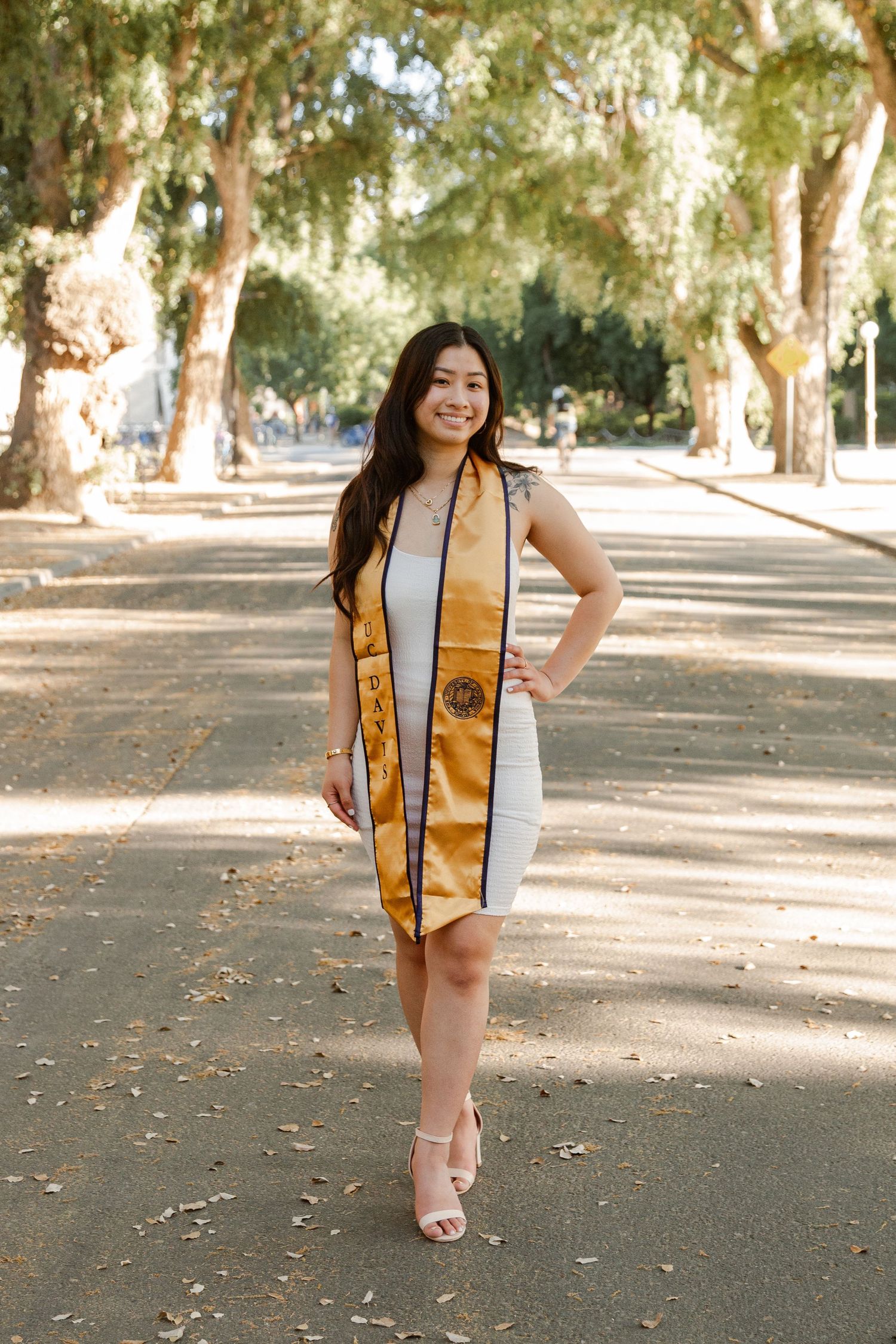 Graduate in white dress and gold sash poses on tree-lined path.