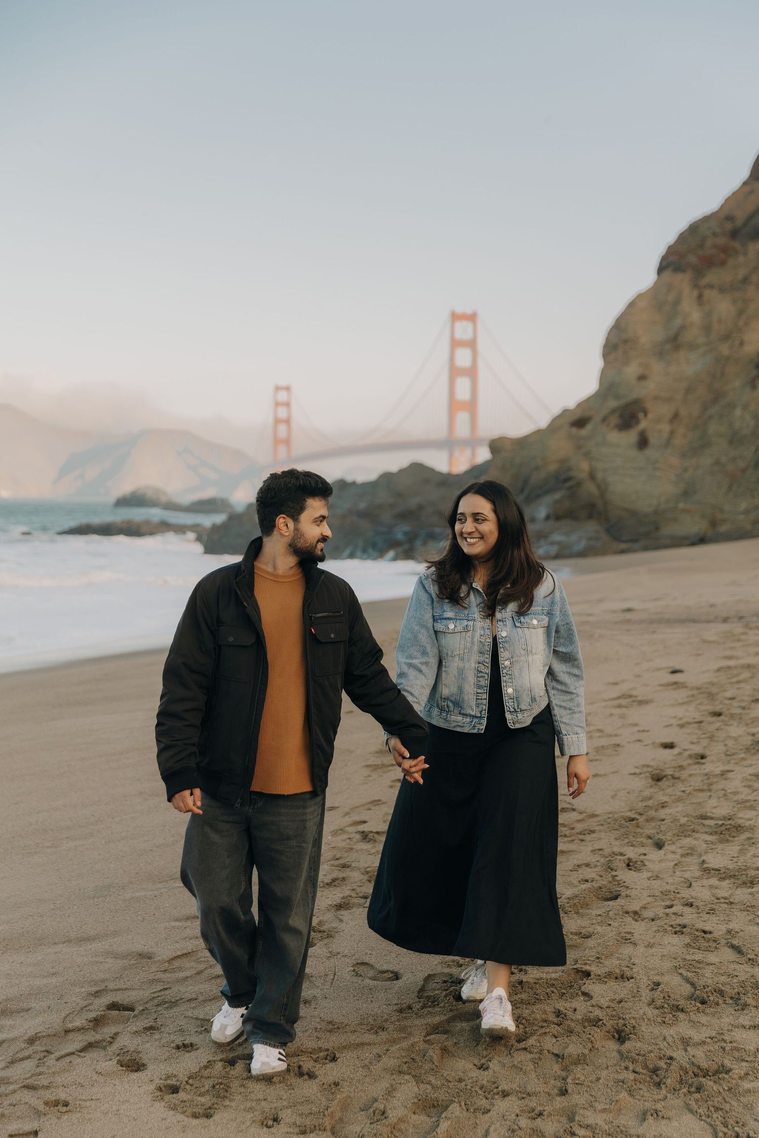 A couple walks along Baker Beach with the iconic Golden Gate Bridge visible in the misty San Francisco background.