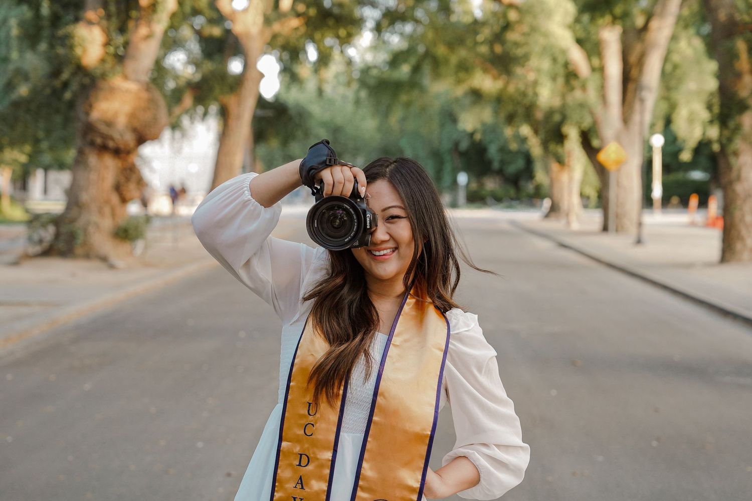 One of Allyson's grad photos from UC Davis — where she holds her camera up and smiles on tree-lined path.