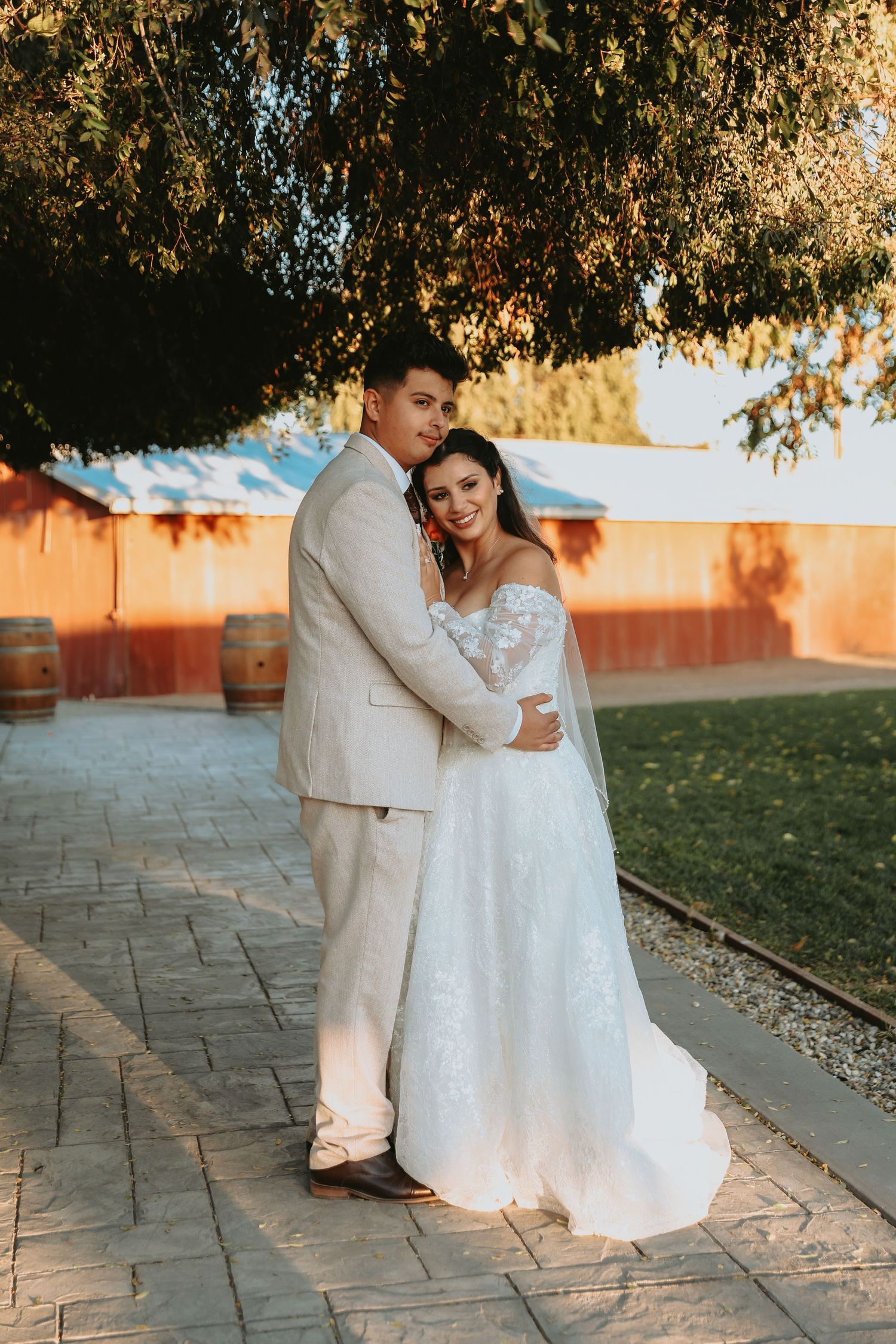 A couple in wedding attire embraces on a stone pathway at sunset with trees and a red wall in the background.