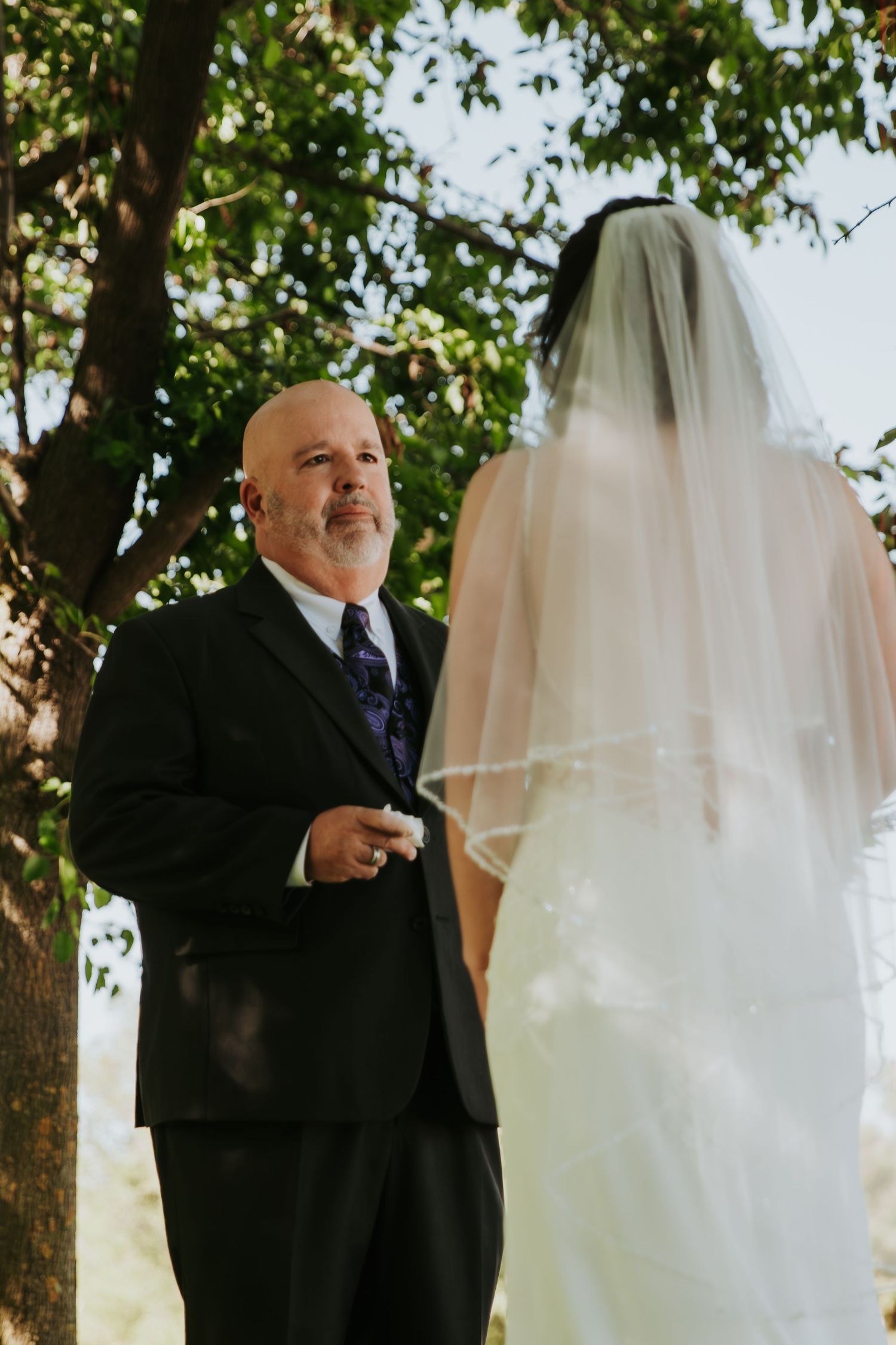 A wedding ceremony outdoors with a bride in white dress and veil standing with someone in a dark suit under trees.