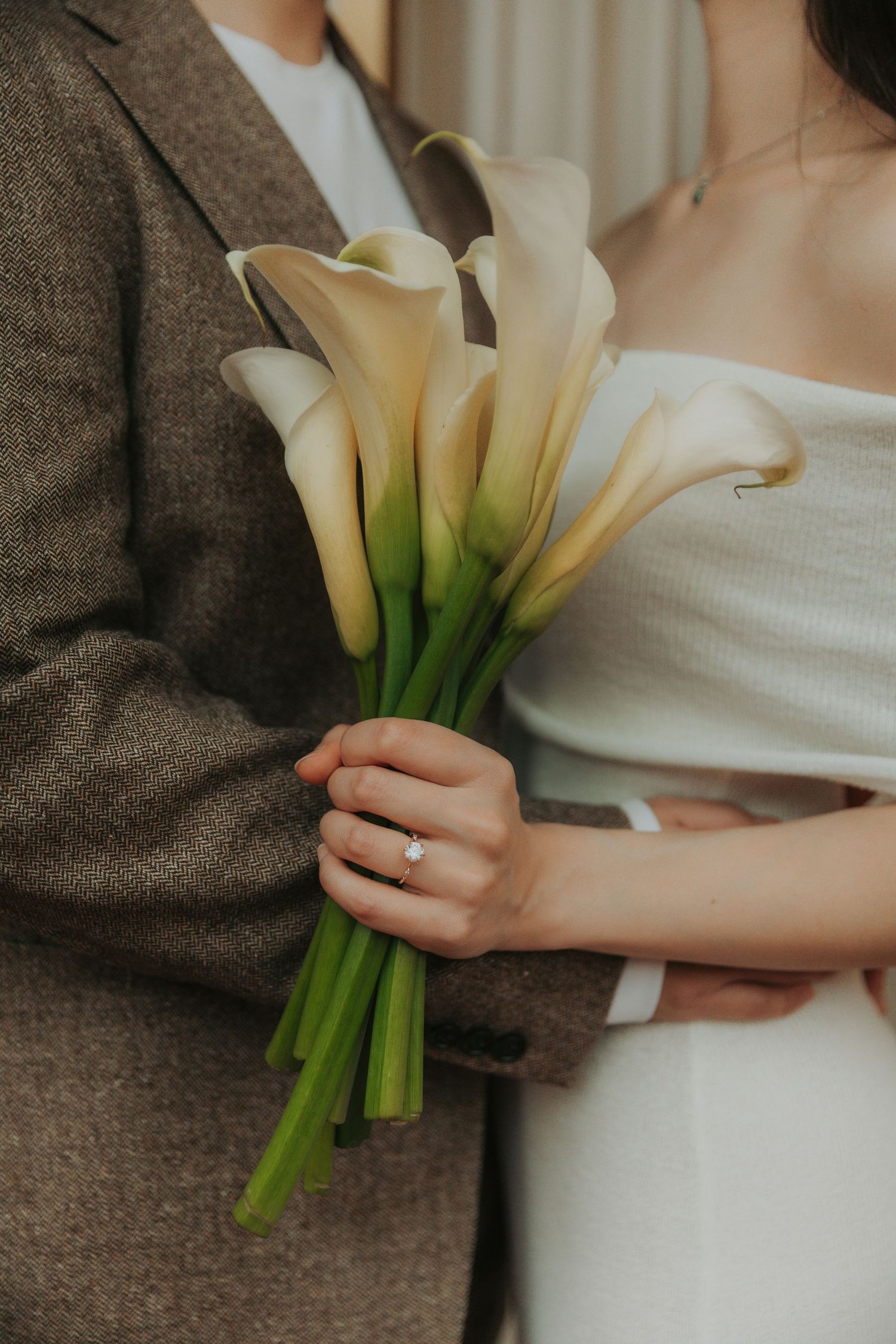 A couple holding a bouquet of white calla lilies together against a neutral background in an intimate setting.