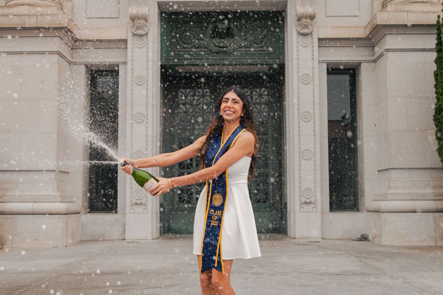 A graduate in a white dress celebrates with champagne spray in front of stone building.