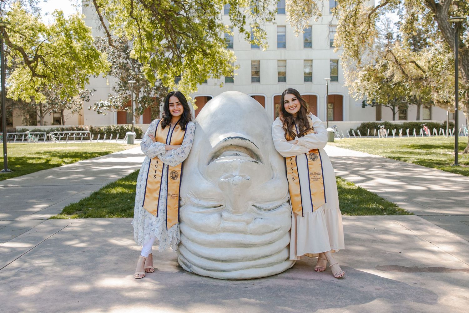 Graduates in light dresses pose with large sculpture head on university campus.