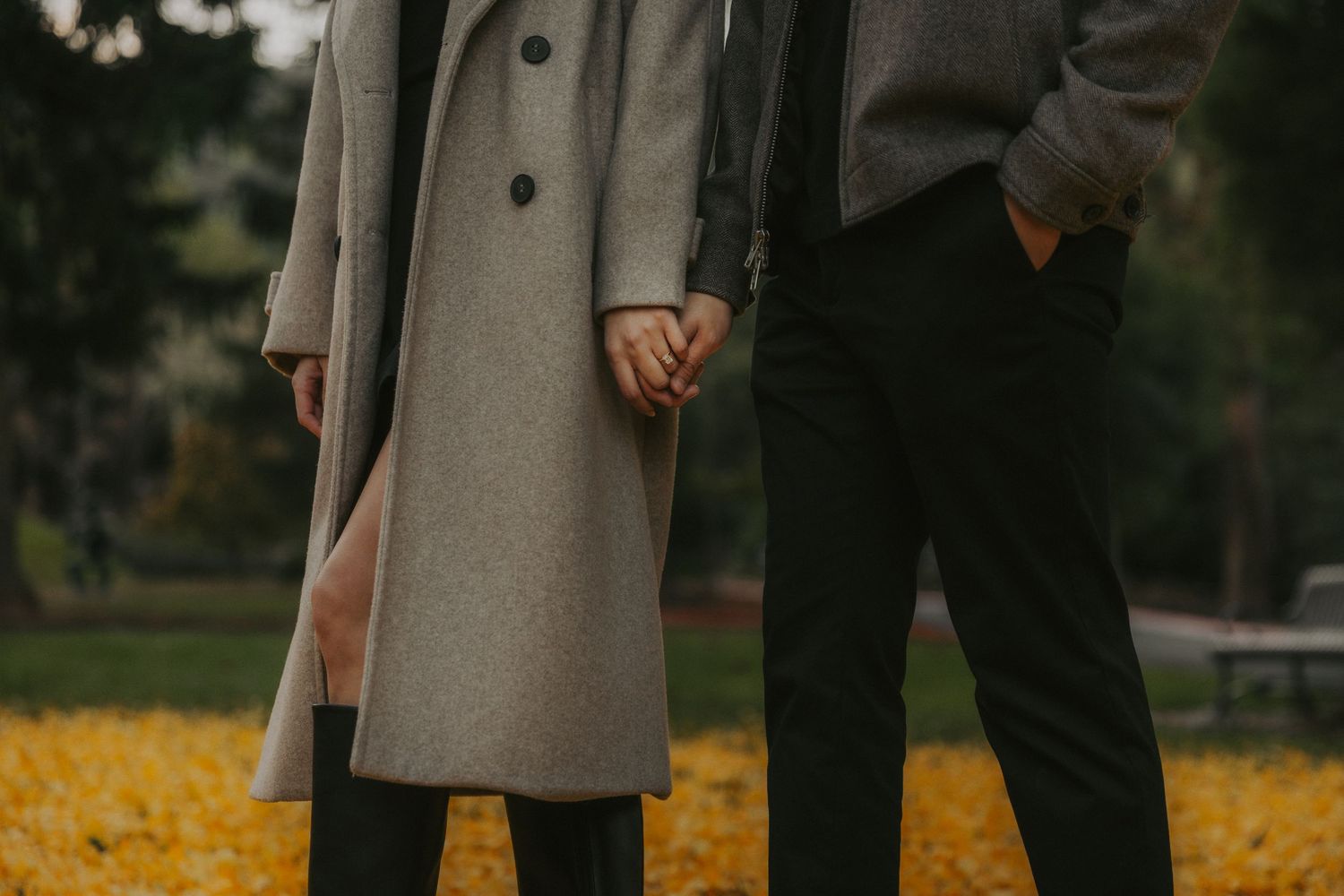 A couple holding hands walks through fallen autumn leaves on a path in a park.