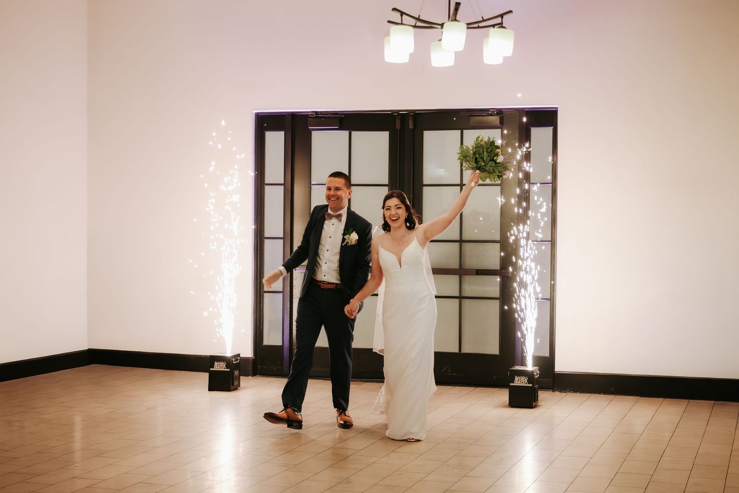A couple makes their grand entrance at a wedding reception through modern black-framed glass doors with string lights.