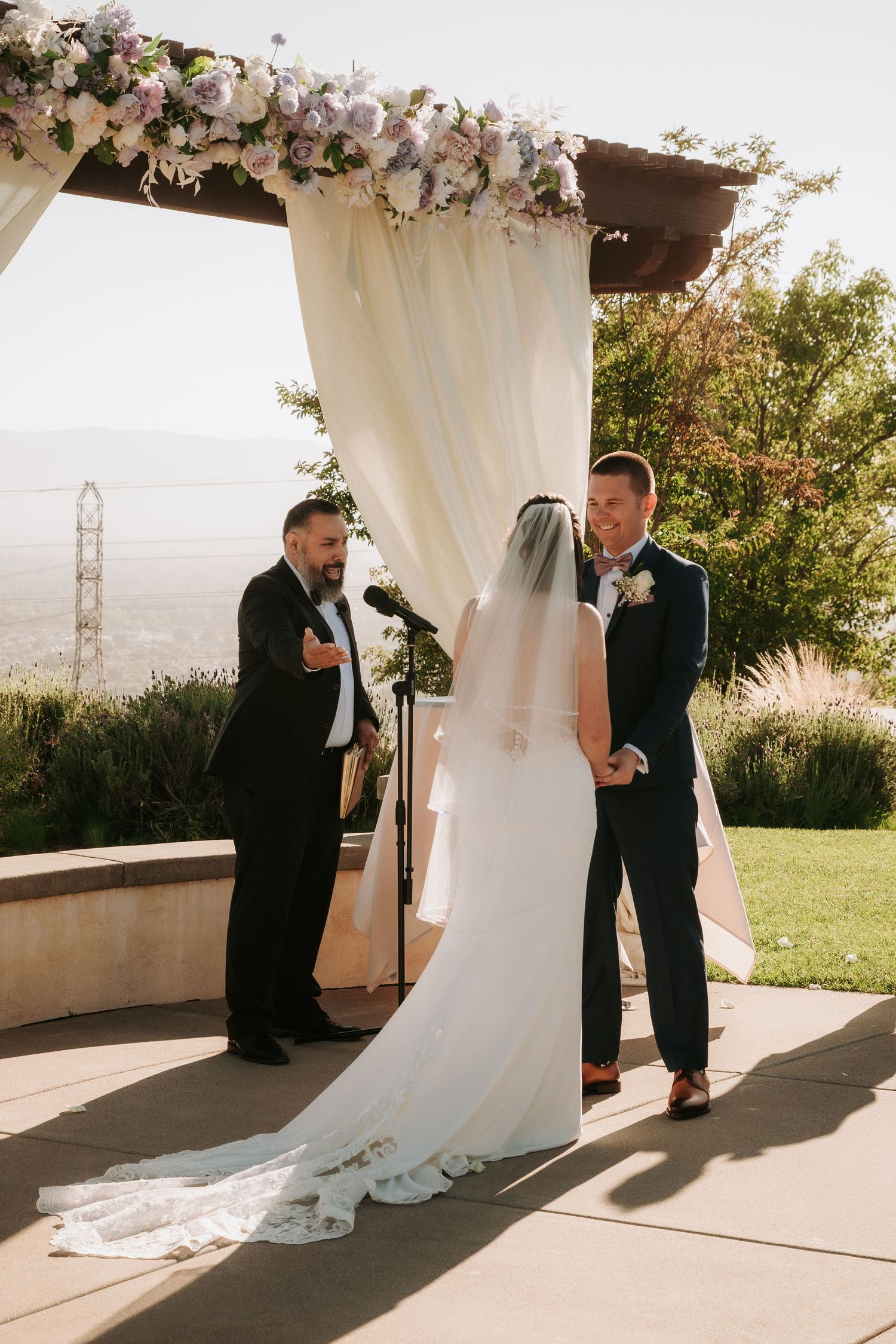 Wedding couple shares first look moment under white floral arch with scenic mountain backdrop.