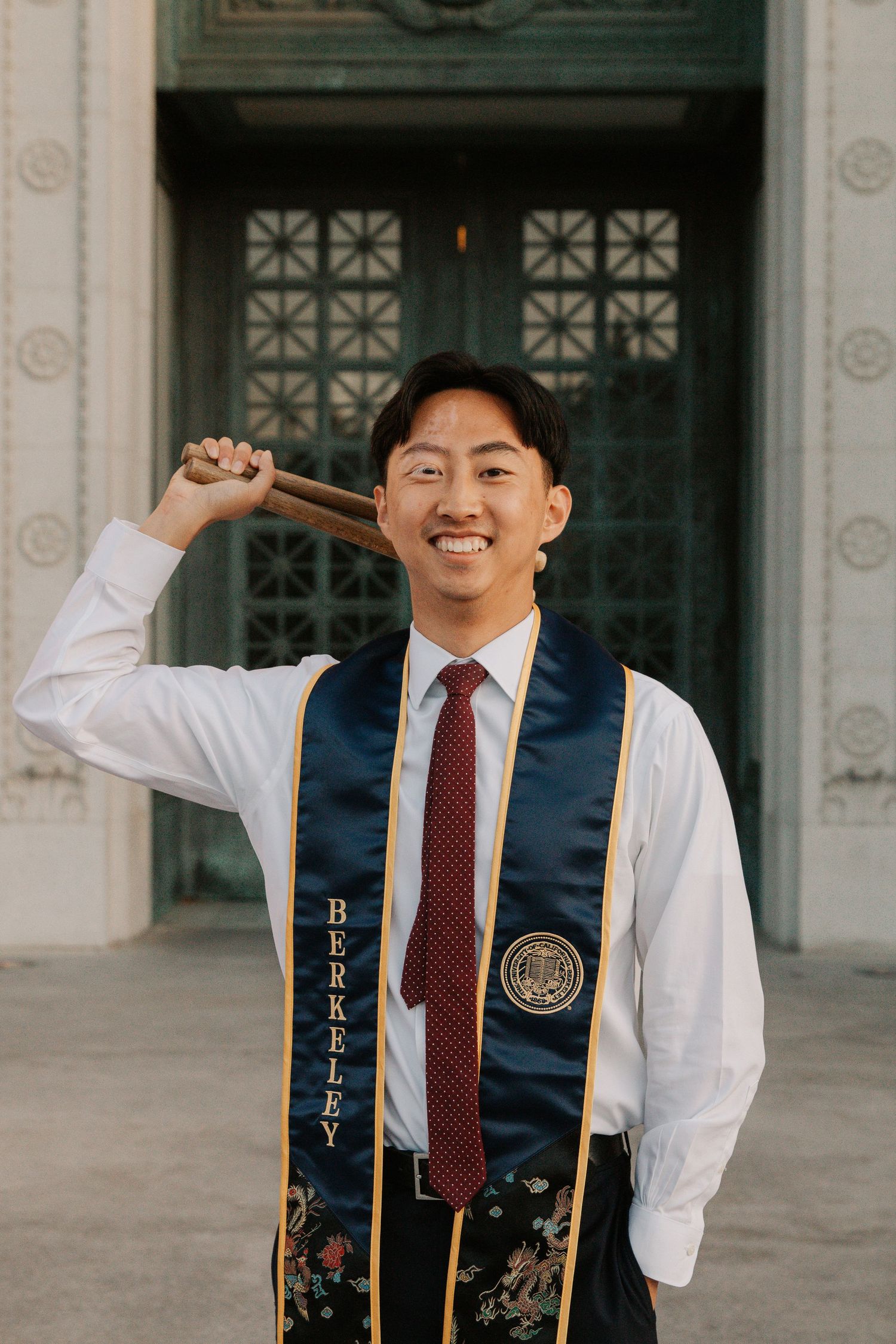 Graduate in formal attire poses with graduation sash by ornate doorway.