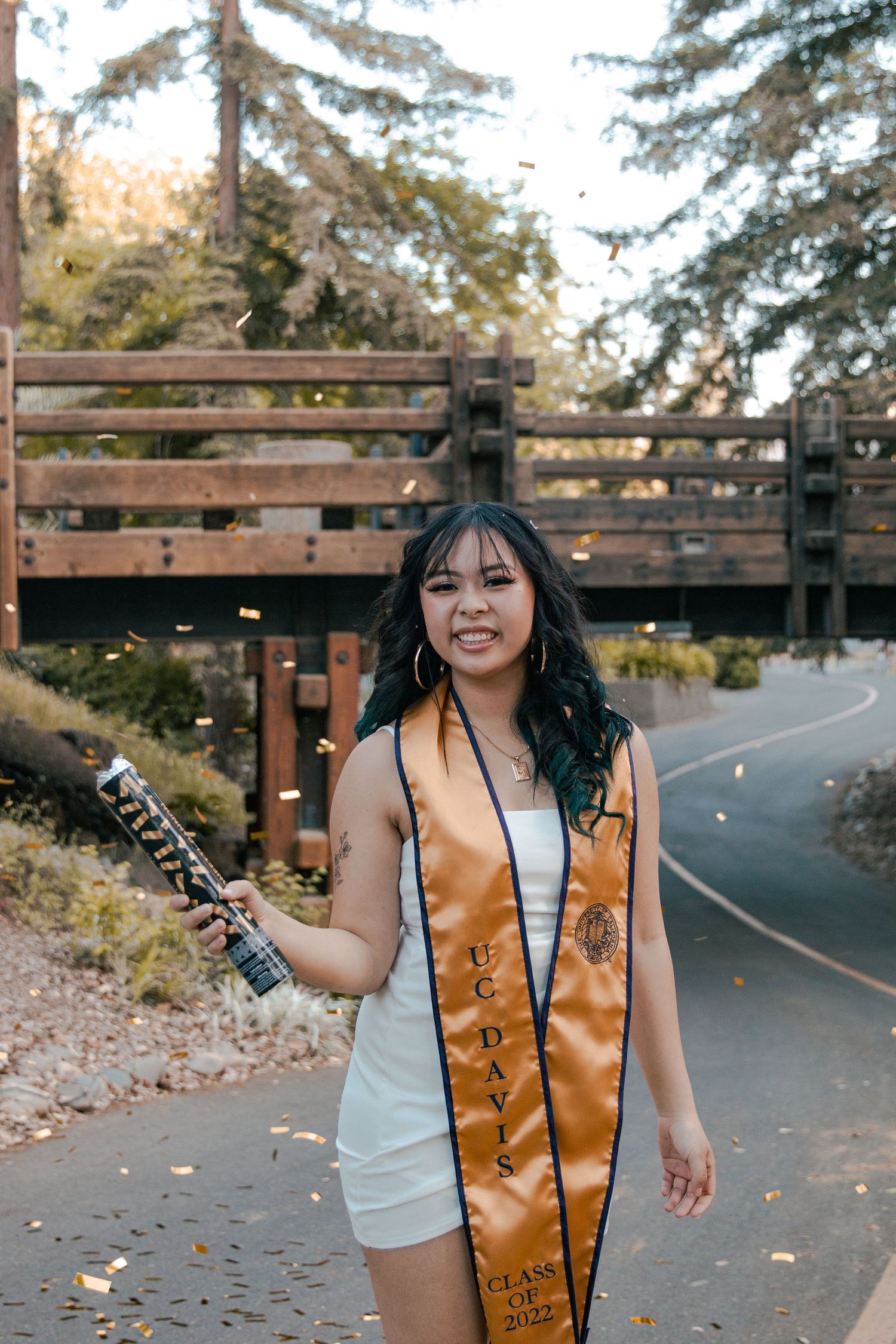 Graduate in white dress poses outdoors with gold graduation stole against wooden bridge background.
