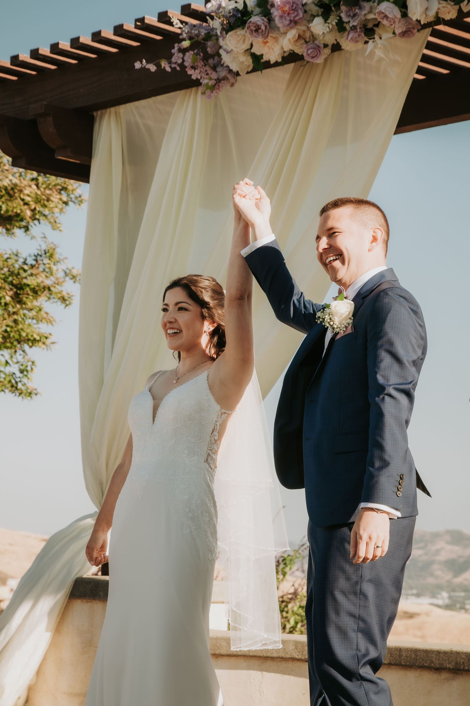 Joyful celebration during outdoor wedding ceremony under floral-adorned pergola with white fabric draping.