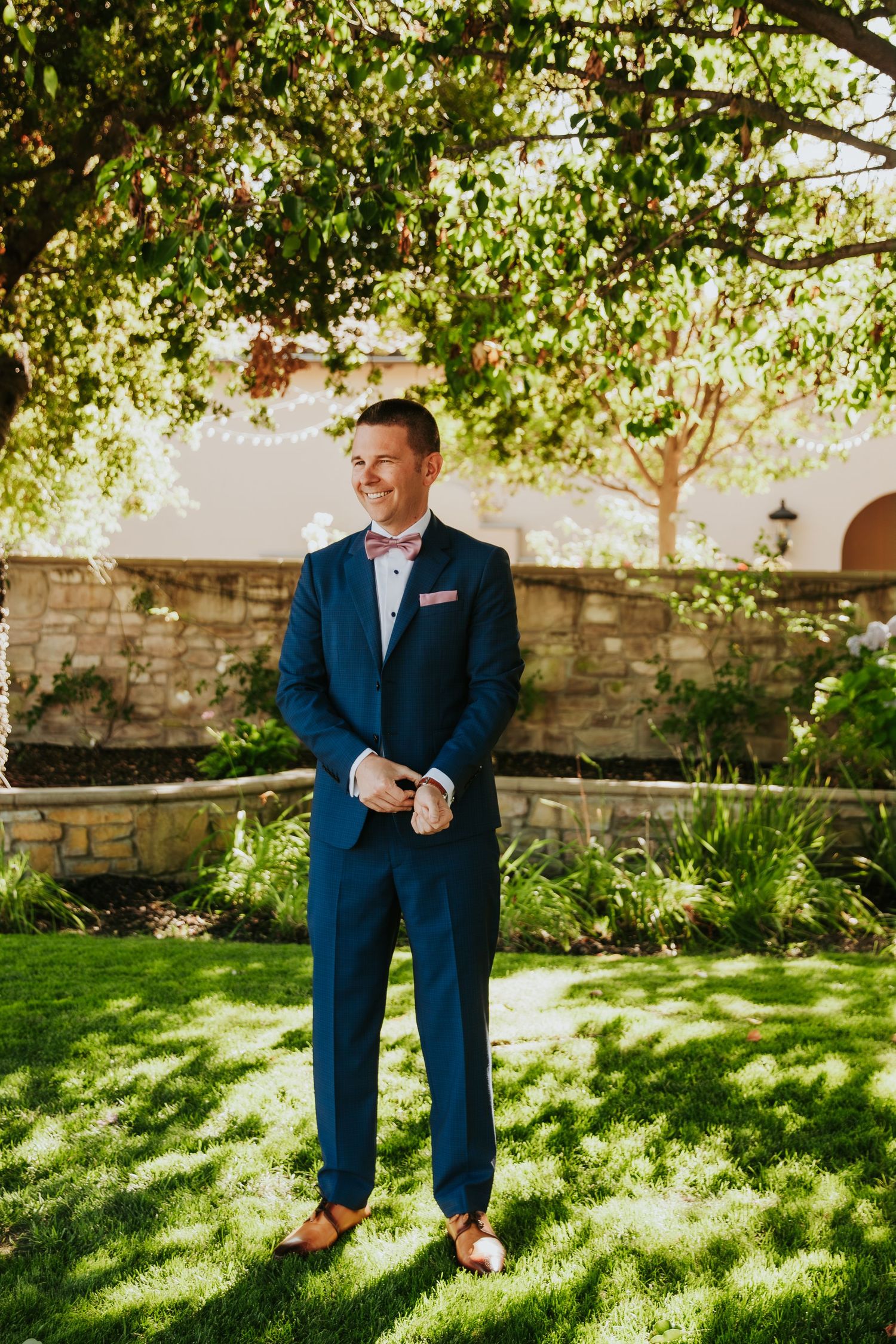 A person in a navy blue suit stands on green grass under dappled sunlight near a stone wall.
